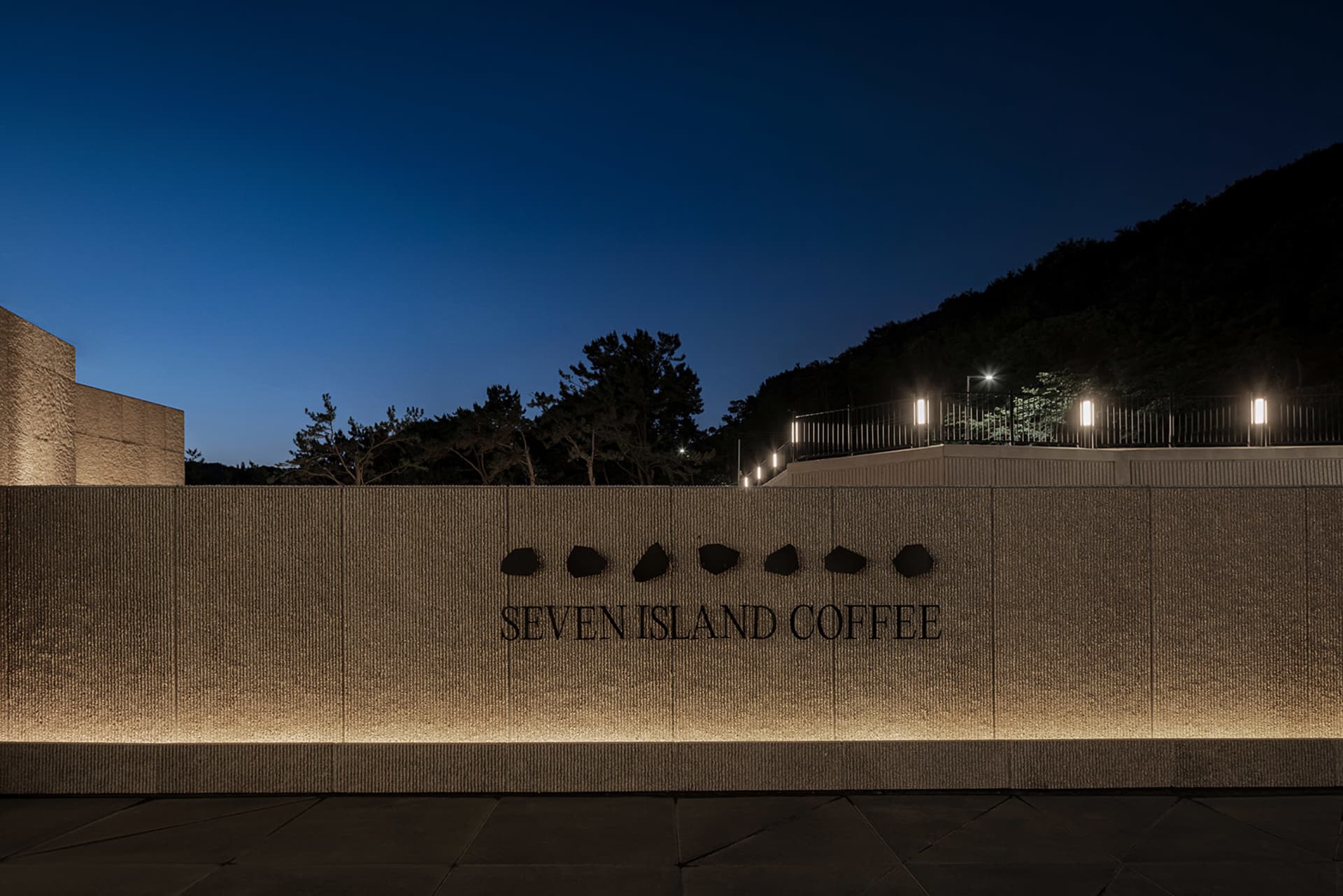 A coffee shop at night with a stone wall featuring embedded rocks and a sign reading Seventen Island Coffee, lit up against the dark sky