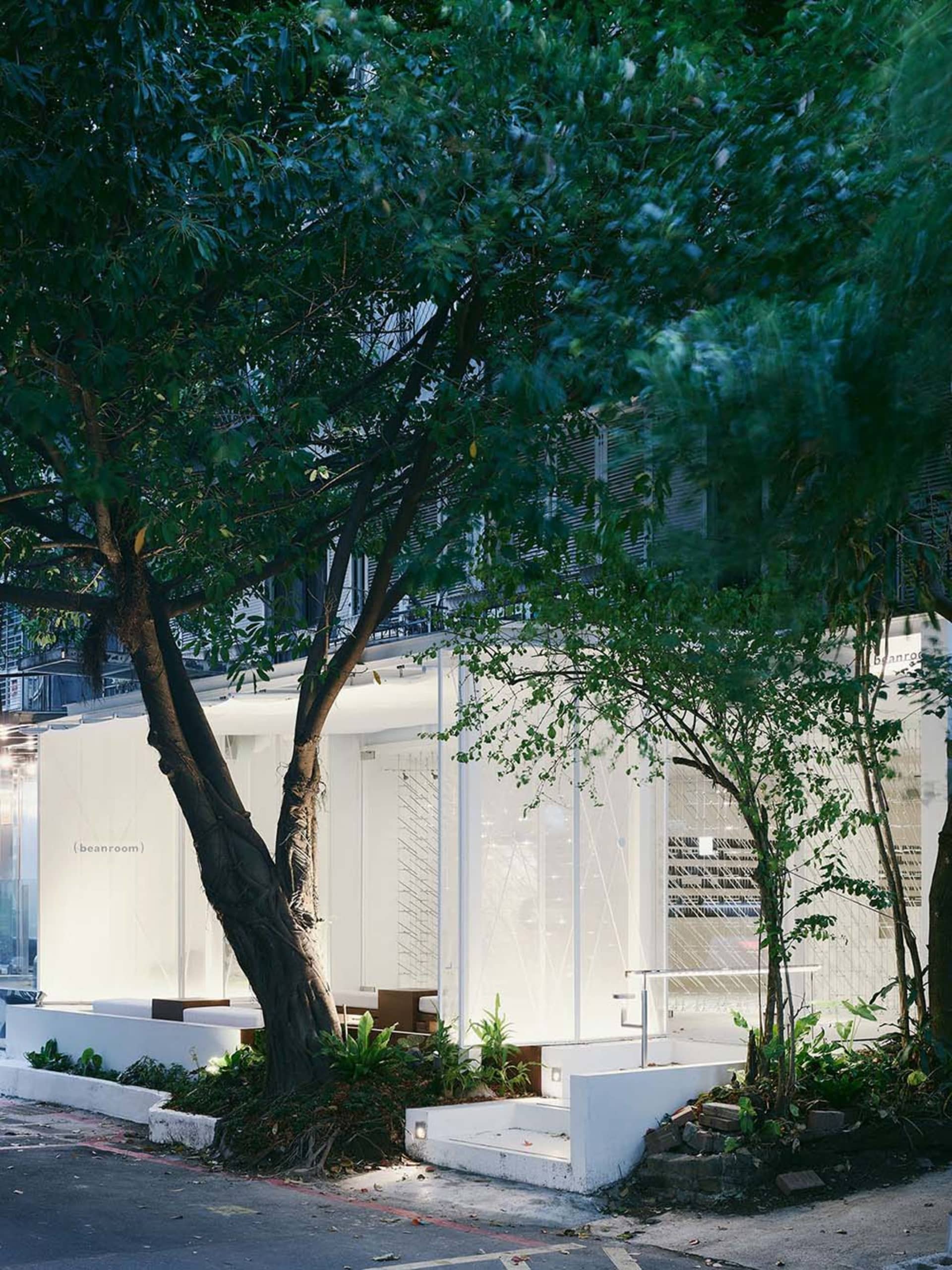 A modern building with a white exterior and glass wall, surrounded by chairs and potted plants, with a tree in the foreground