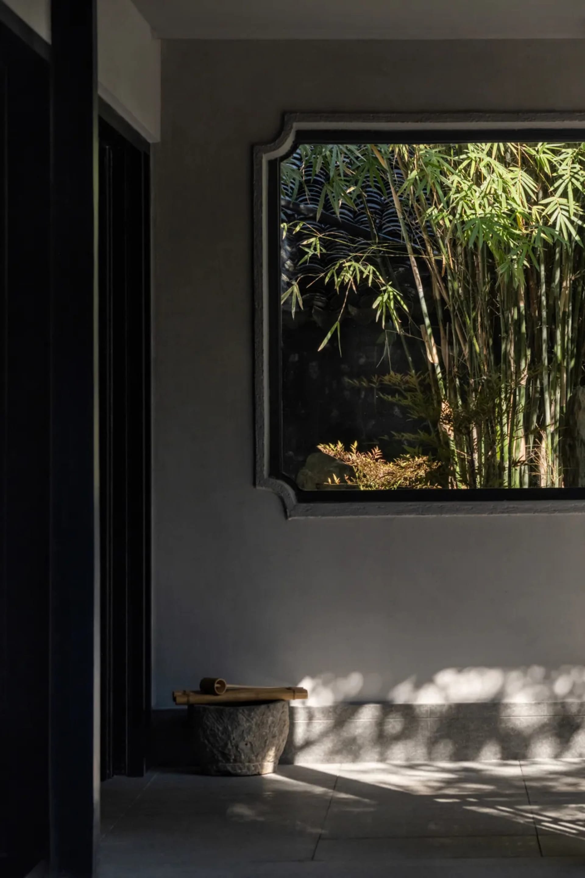 A serene Asian-style room with a gray wall, a window with bamboo plants outside, and two potted plants in front of the window