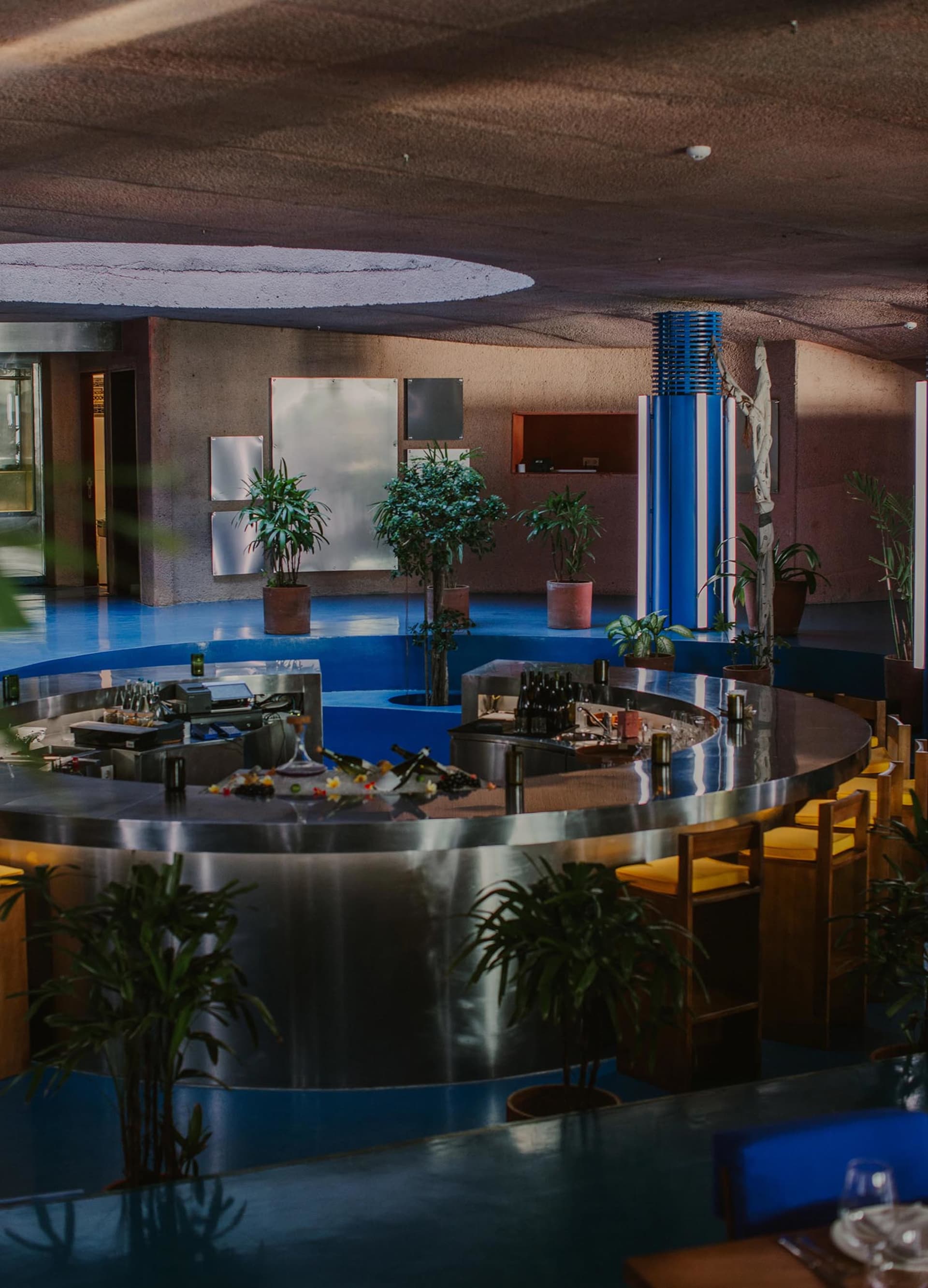 A restaurant interior with a circular bar surrounded by chairs and potted plants, bottles on the countertop, and a welcoming atmosphere