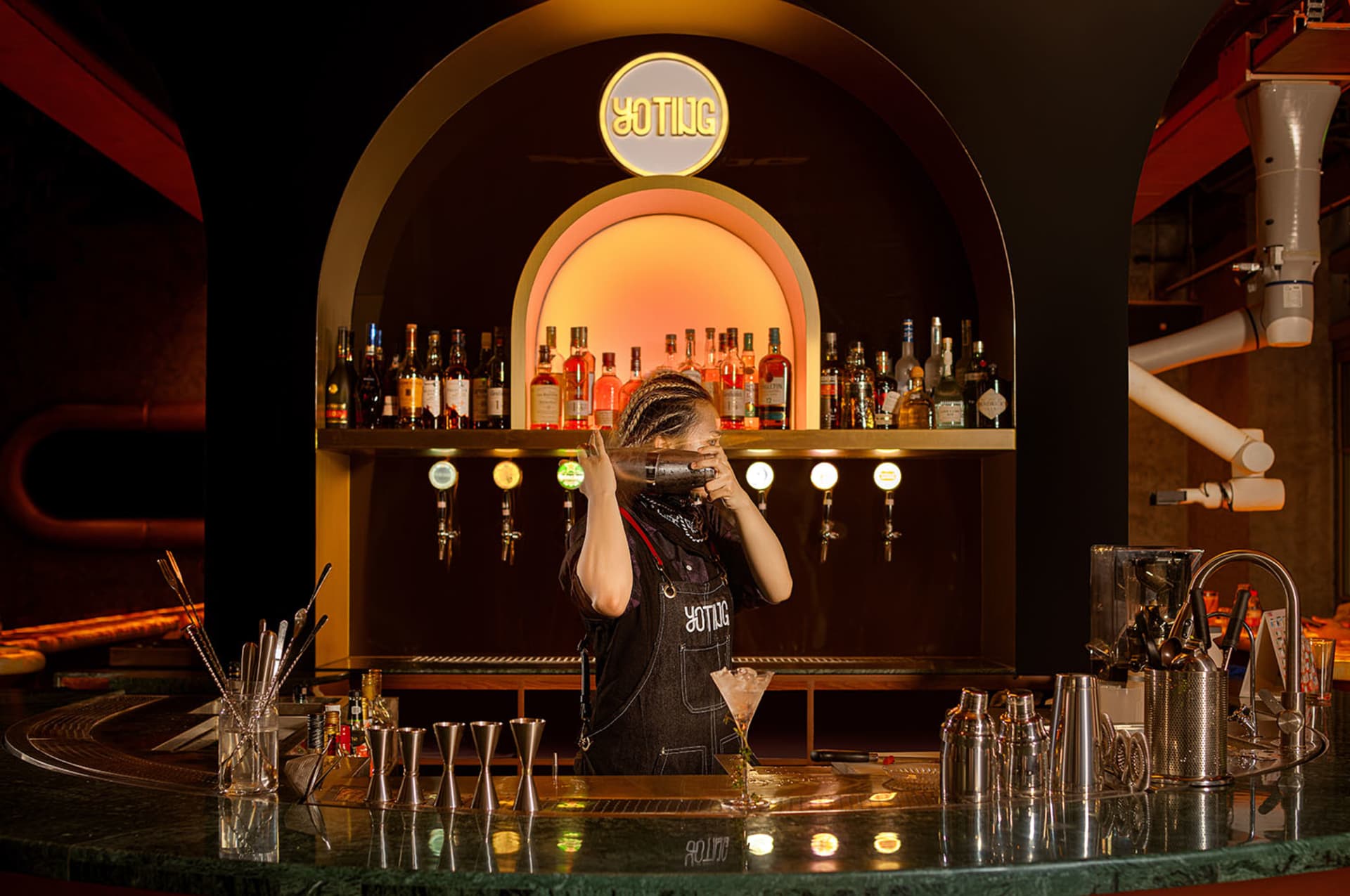 A lively bar scene with a woman behind the counter, surrounded by bottles, cups, and wine glasses