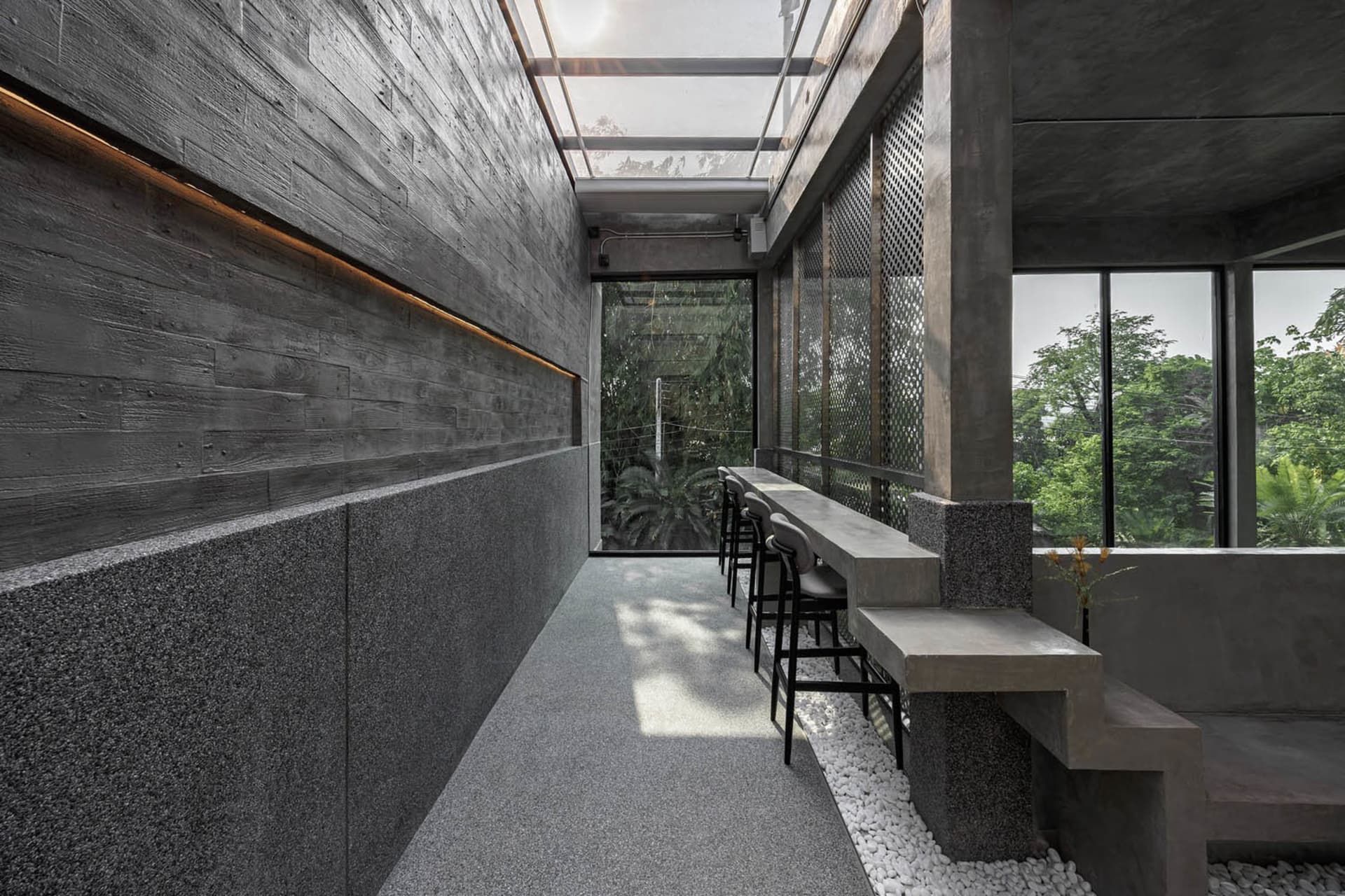 A modern restaurant with a large dining table, concrete walls, a glass ceiling, and potted plants, creating an inviting atmosphere for socializing and dining