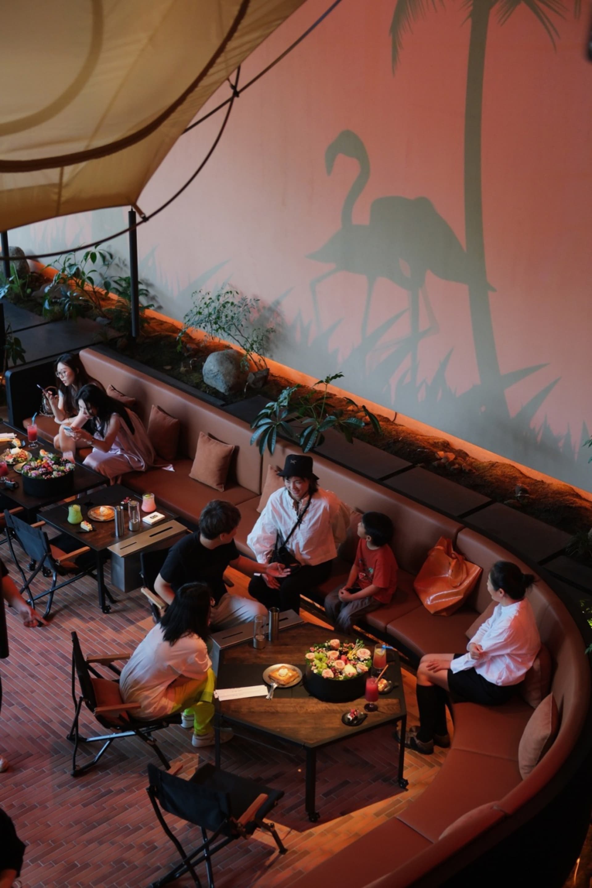A warm-toned restaurant interior with couches, chairs, tables, and potted plants, featuring people enjoying meals and conversation