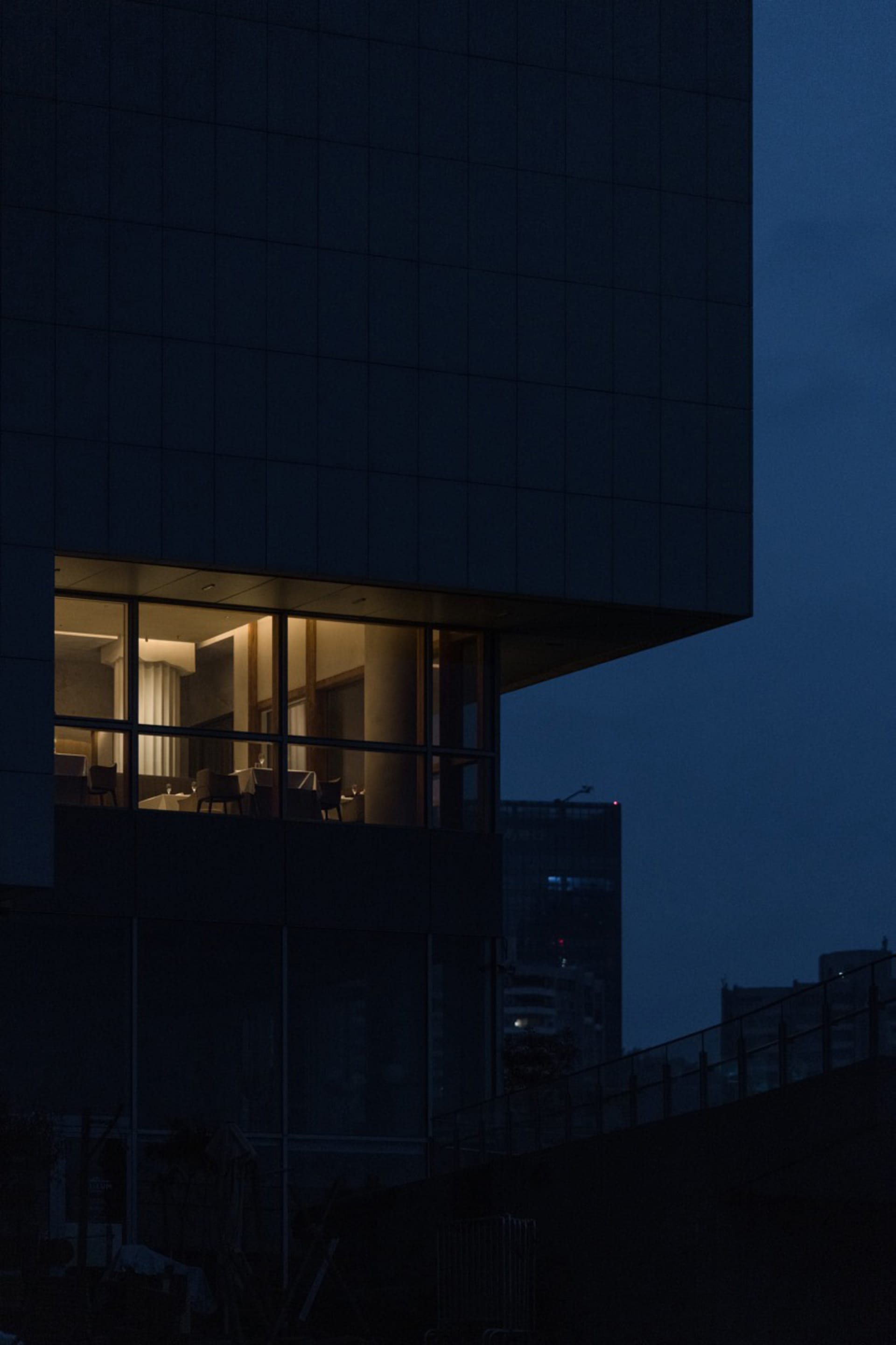 A modern high-rise building at night with a glass structure, multiple windows, and well-lit interior, featuring chairs and a sophisticated atmosphere