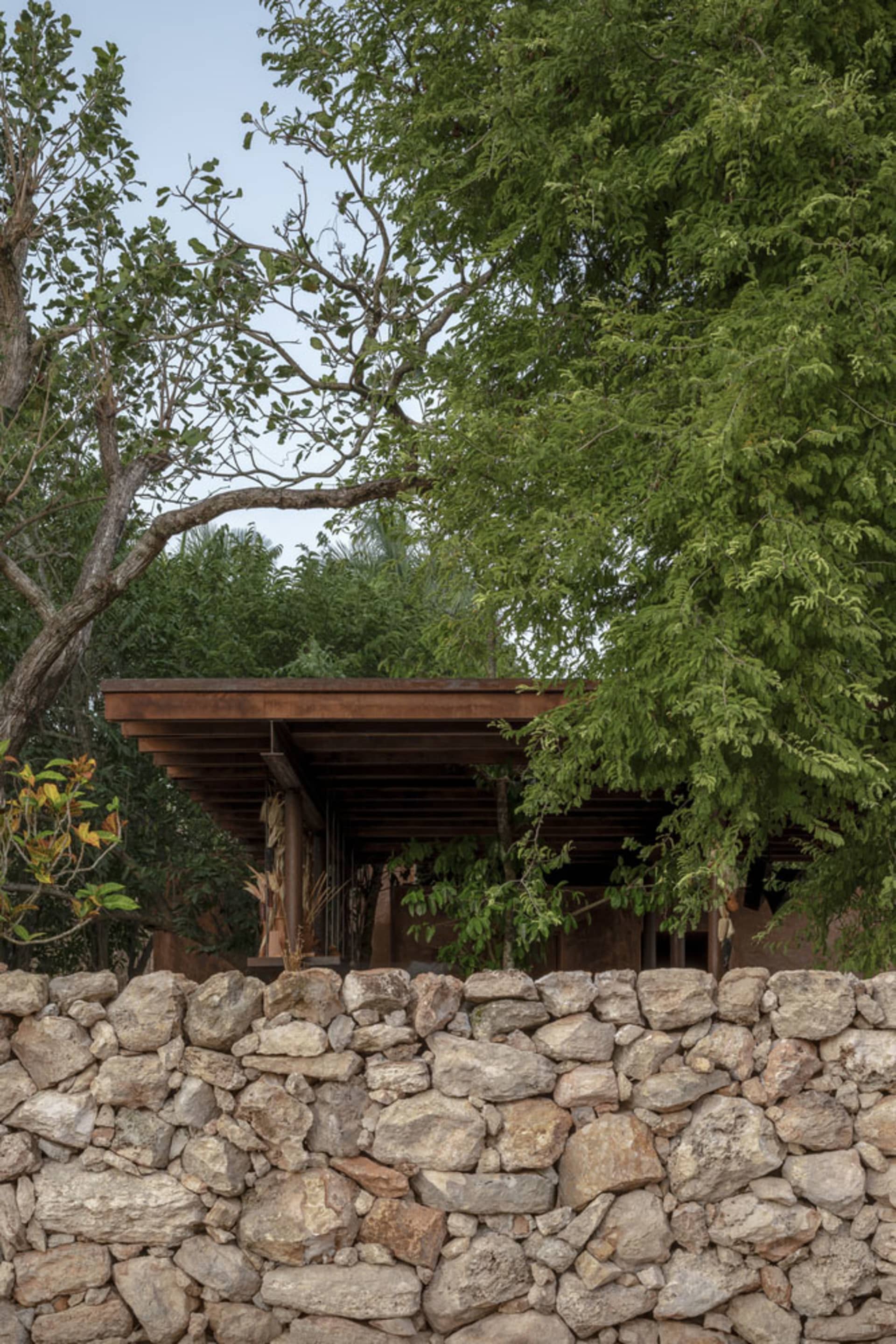 A rustic house with a stone wall in front, surrounded by trees, featuring a wooden structure and brick exterior