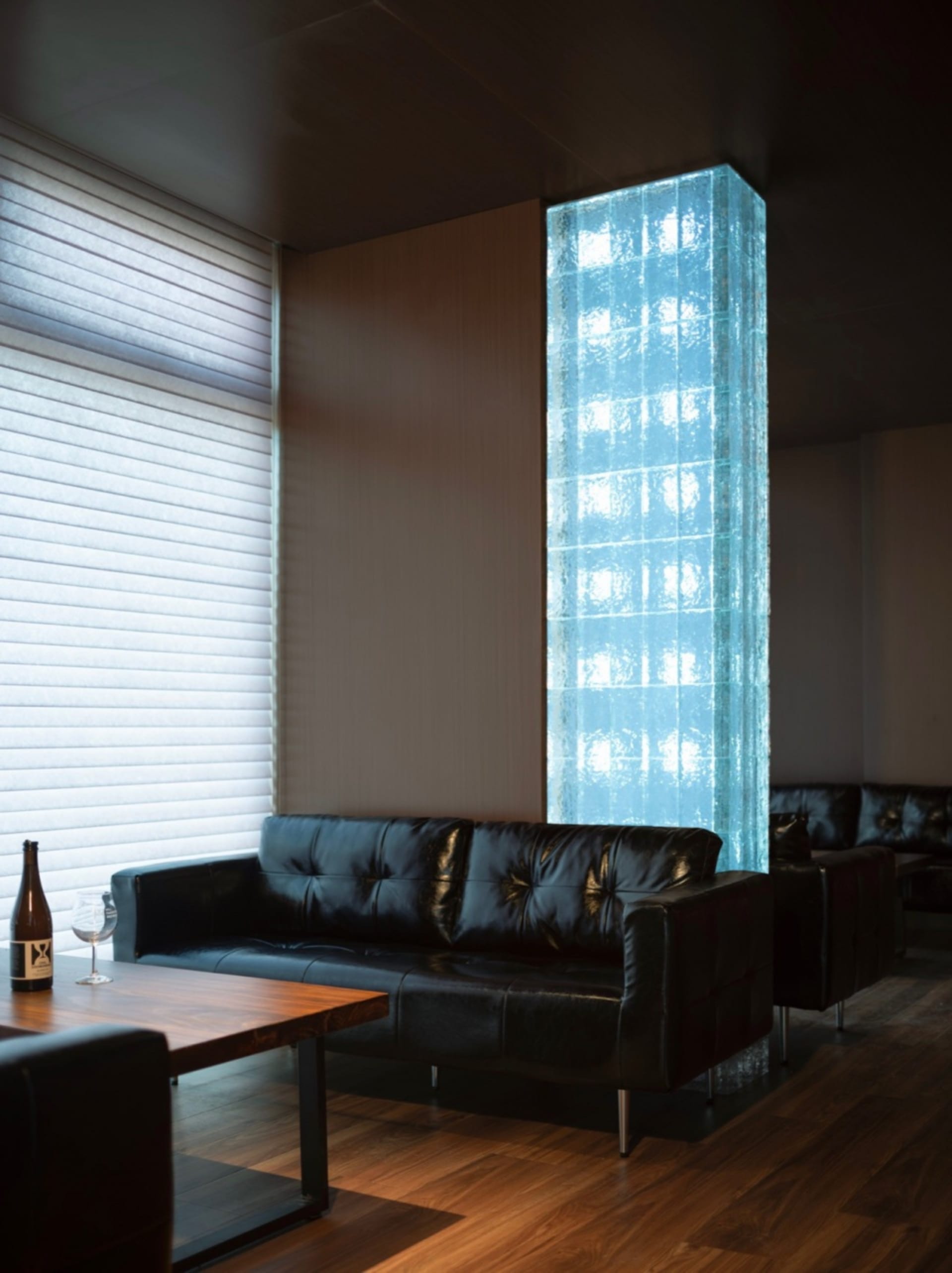Contemporary living space with a black leather couch, wooden table, and a large glass wall with a blue tint, featuring two chairs and a sophisticated design
