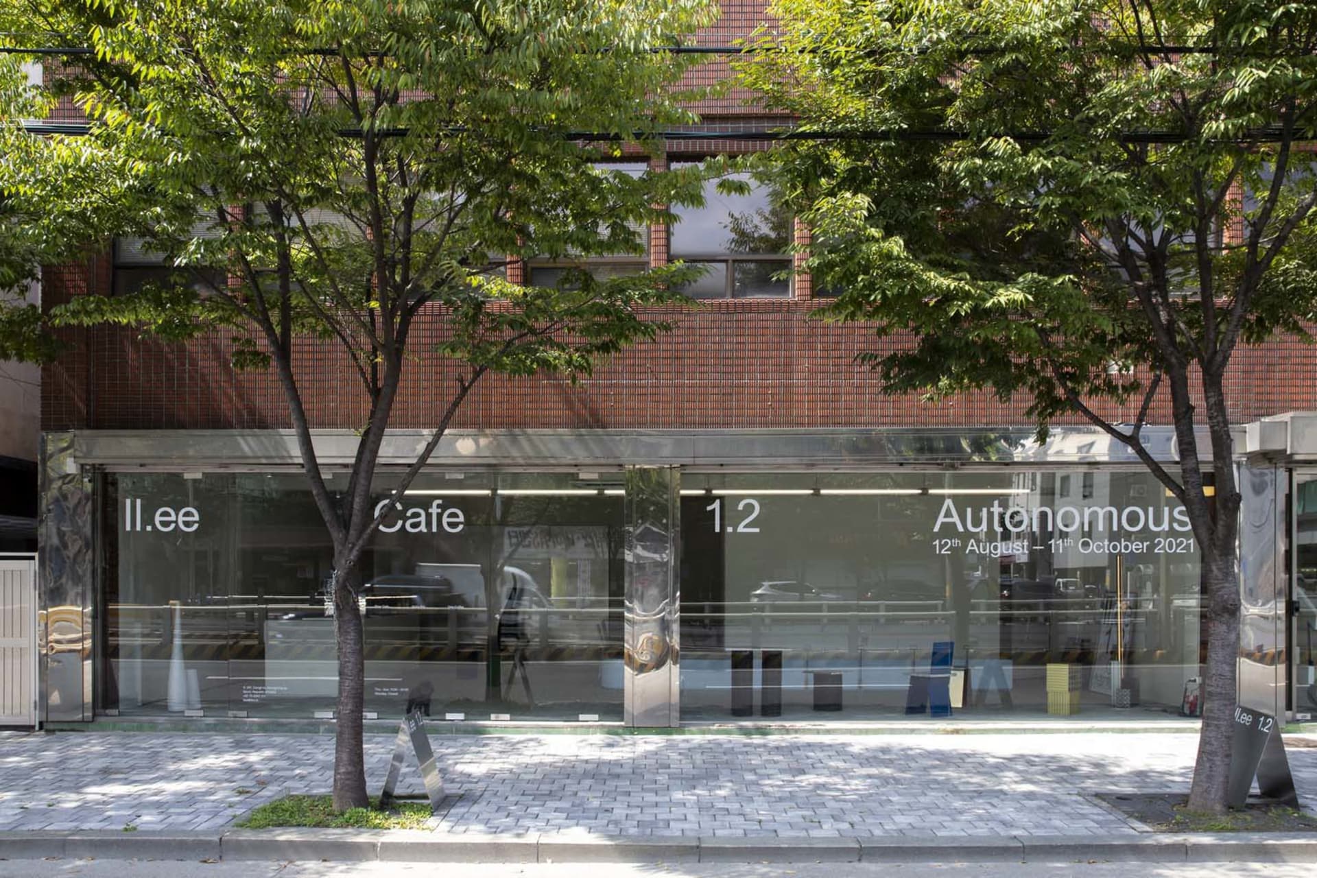 一棟現代咖啡廳建築，擁有磚牆外觀、大片窗戶上寫著「Cafe」，外頭停放數輛汽車，周圍環繞樹木與城市氛圍