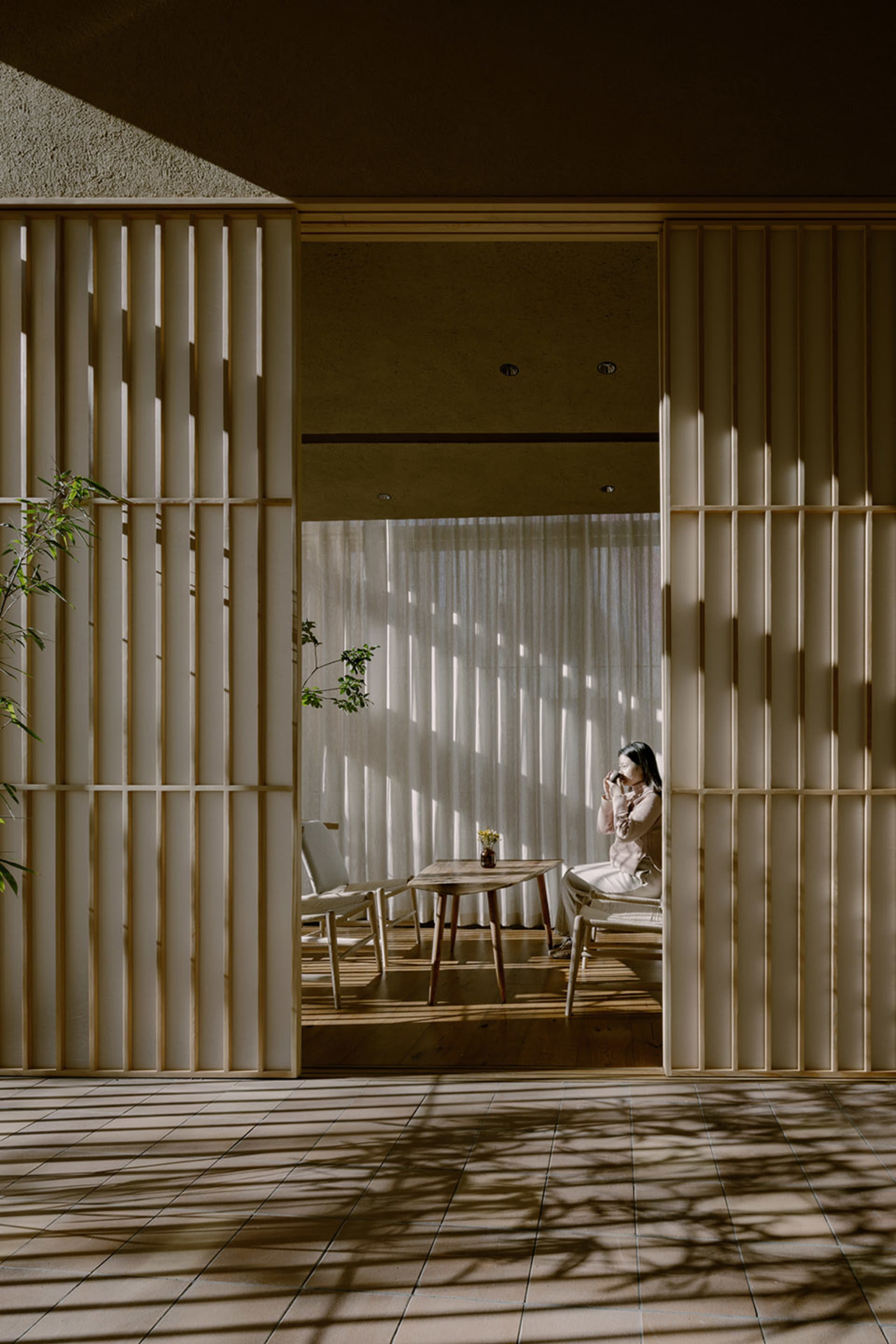 A well-lit indoor living space with a woman sitting on a chair near a dining table, surrounded by wooden furniture and a vase, creating a cozy atmosphere