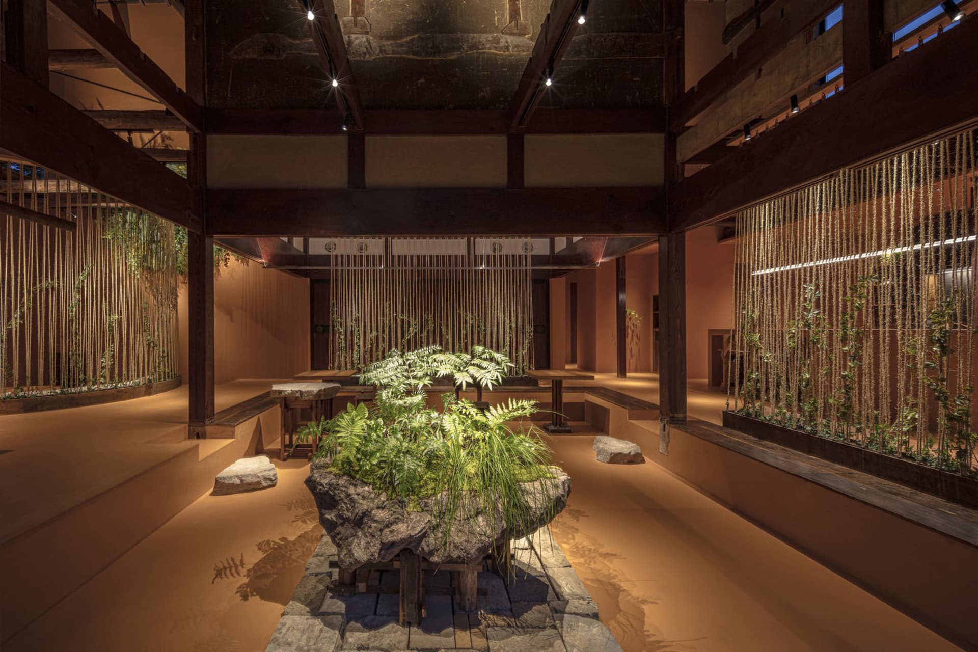 A serene indoor space with a large rock, plants, and wooden floor, featuring multiple benches for seating and a calm atmosphere