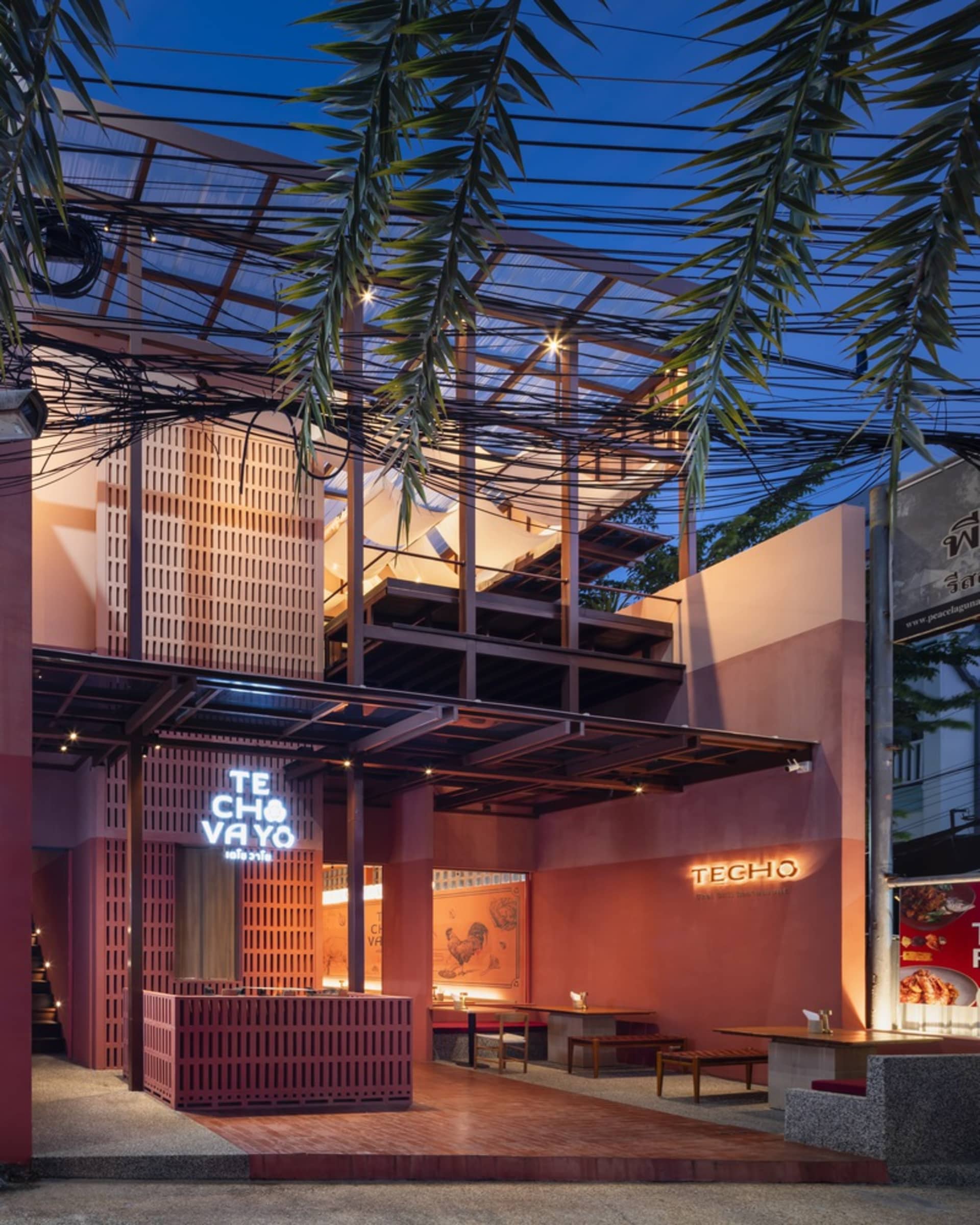 Modern open-air restaurant with a dining area, bar counter, and potted plants, featuring red walls and a warm atmosphere