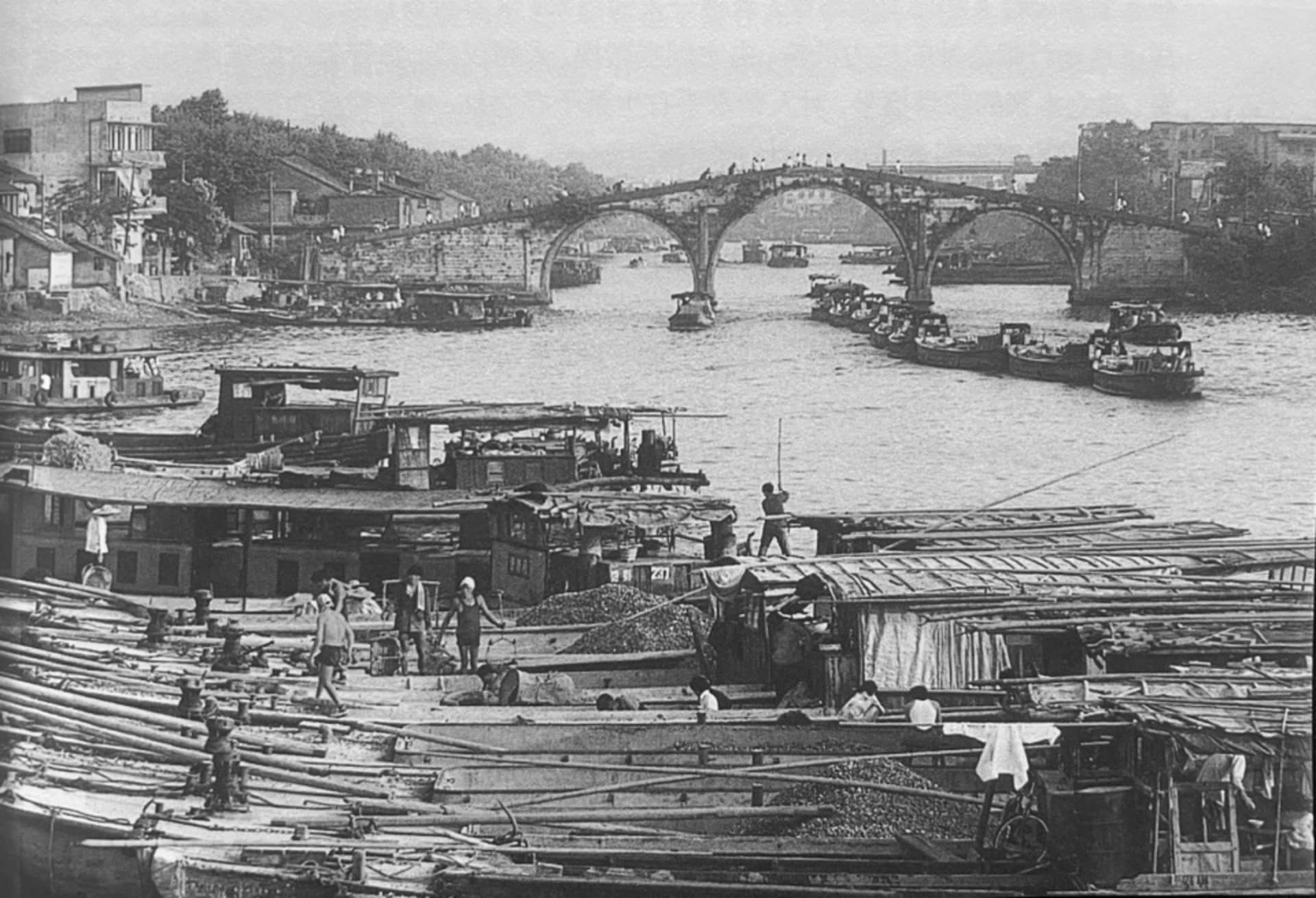 A bustling harbor scene with boats and people in a historic city, with a large bridge over a river
