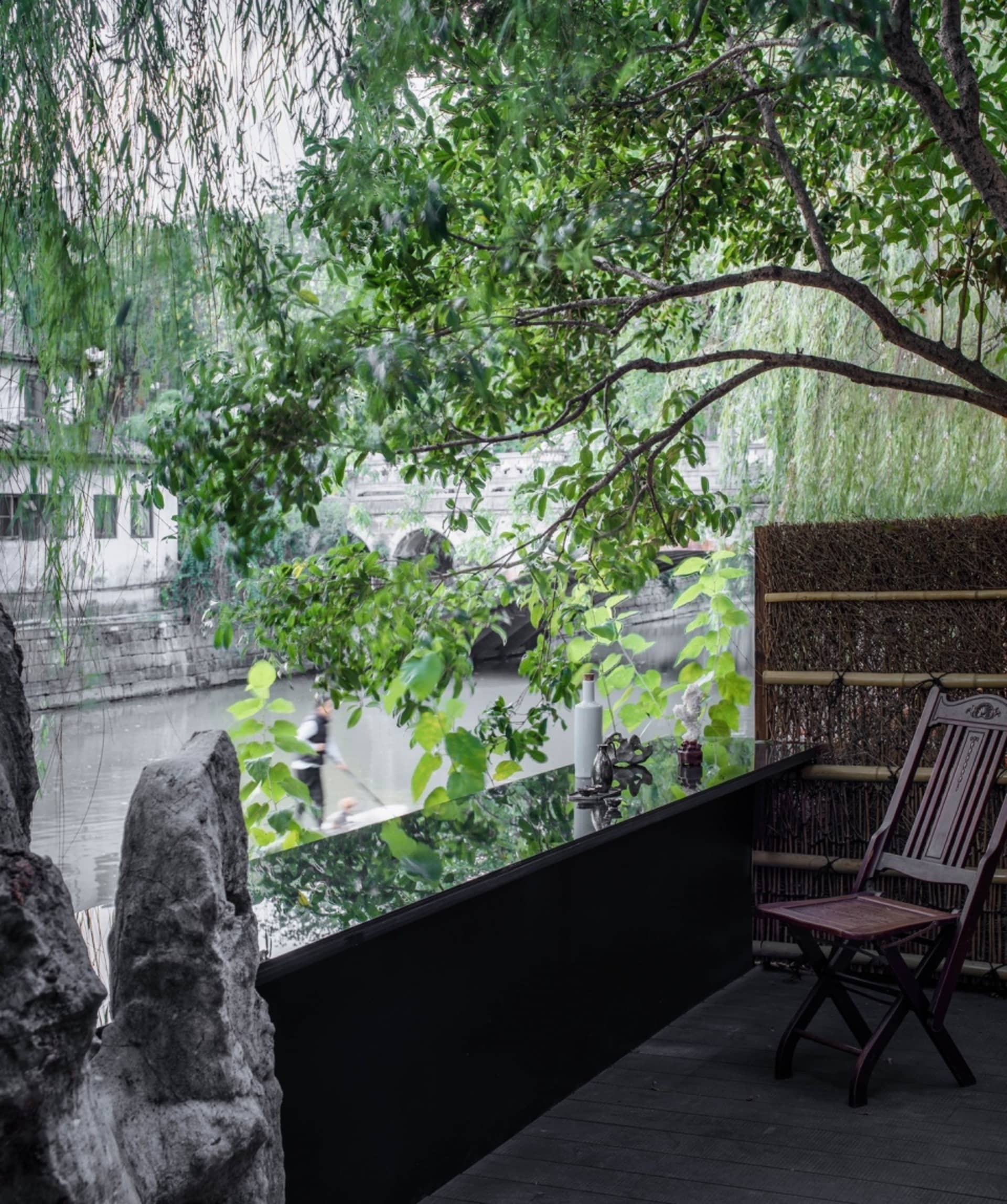 A serene outdoor patio area with a wooden chair on a stone surface near a tree, surrounded by other chairs and lush greenery