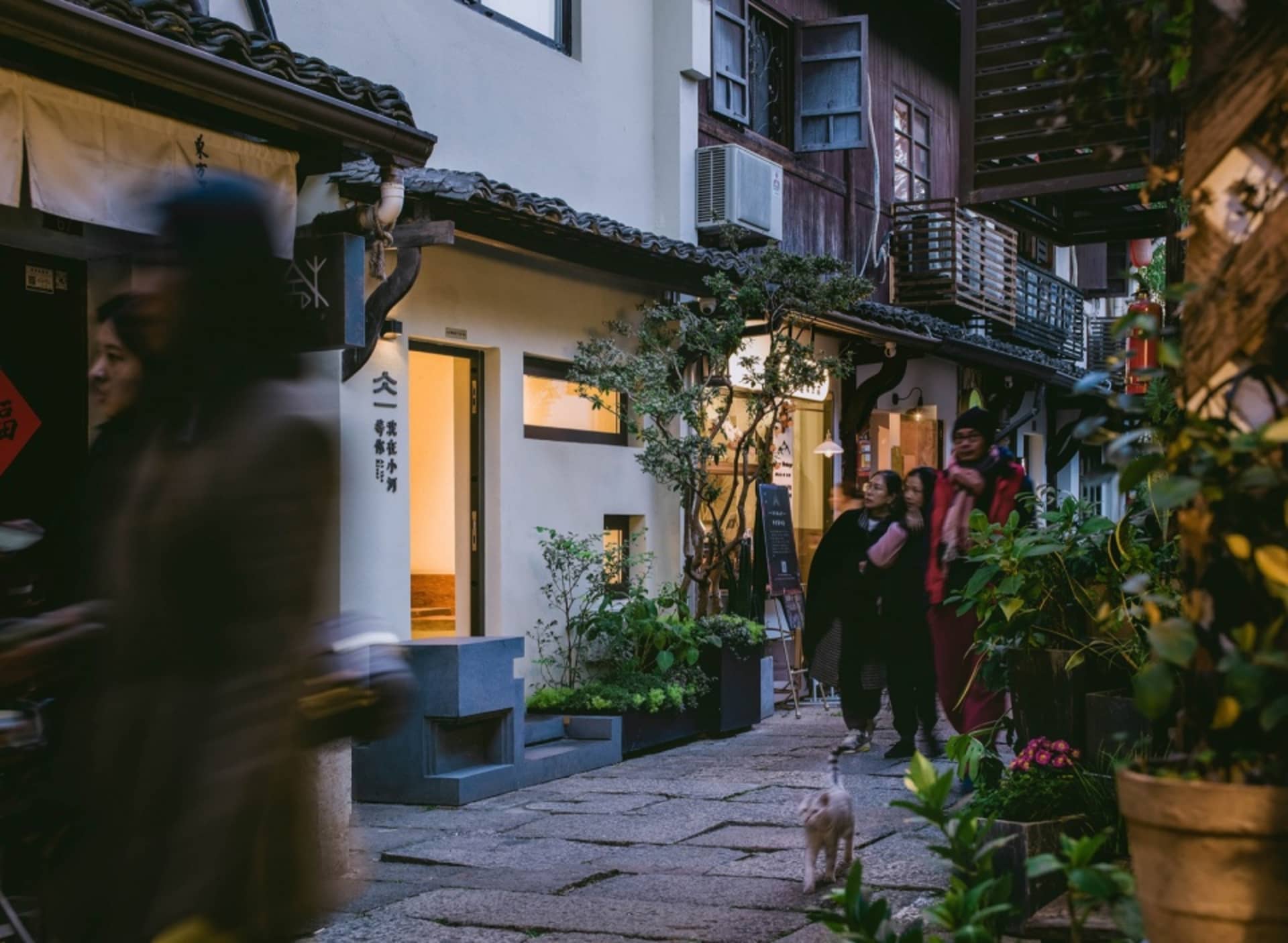 A bustling Asian street scene with a mix of architectural styles, lined with plants and potted flowers, featuring people and a small white dog walking down the sidewalk
