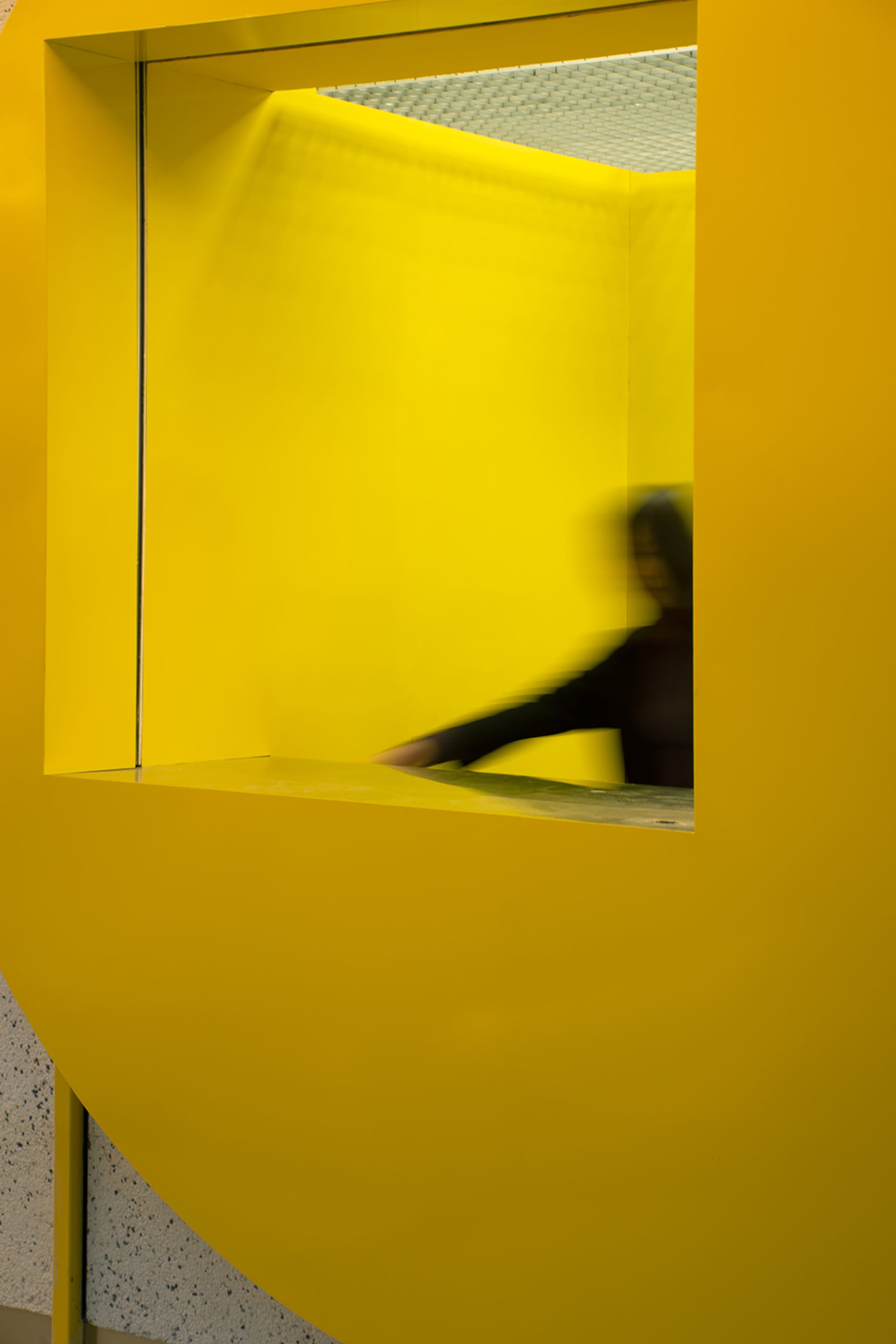 A woman stands in front of a yellow wall, possibly interacting with a door or object, in a residential or public building with a distinctive design element