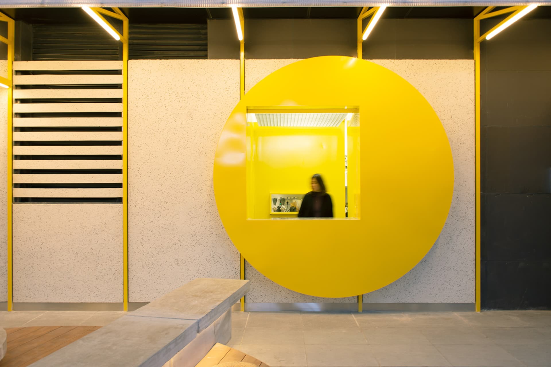 A modern interior space with a large yellow circle on the wall, possibly a window or doorway, with two people looking inside, set in a minimalist design