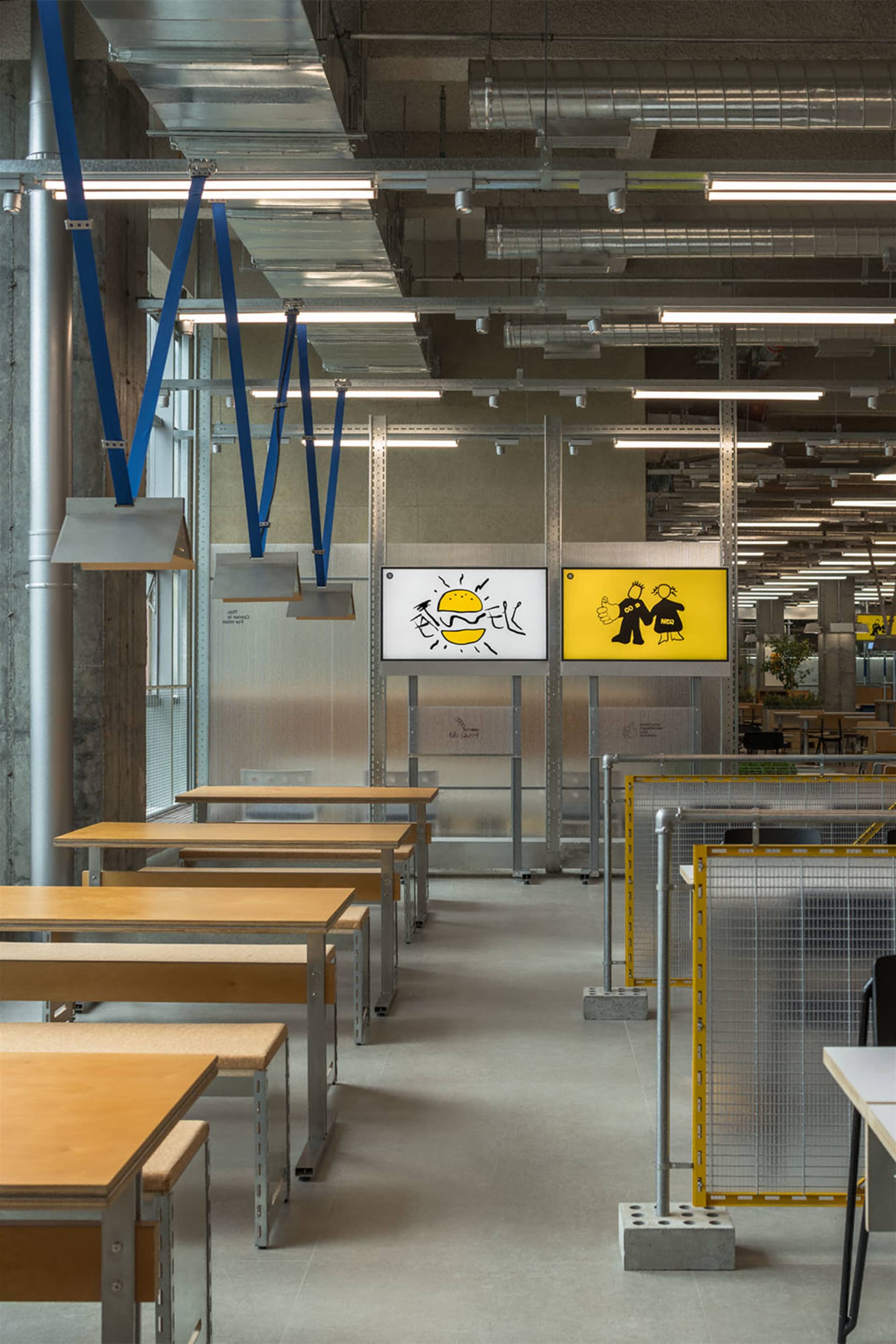 A modern office space with multiple tables and chairs, featuring a mix of wood and metal elements, and yellow signs on some tables, set in a well-lit atmosphere