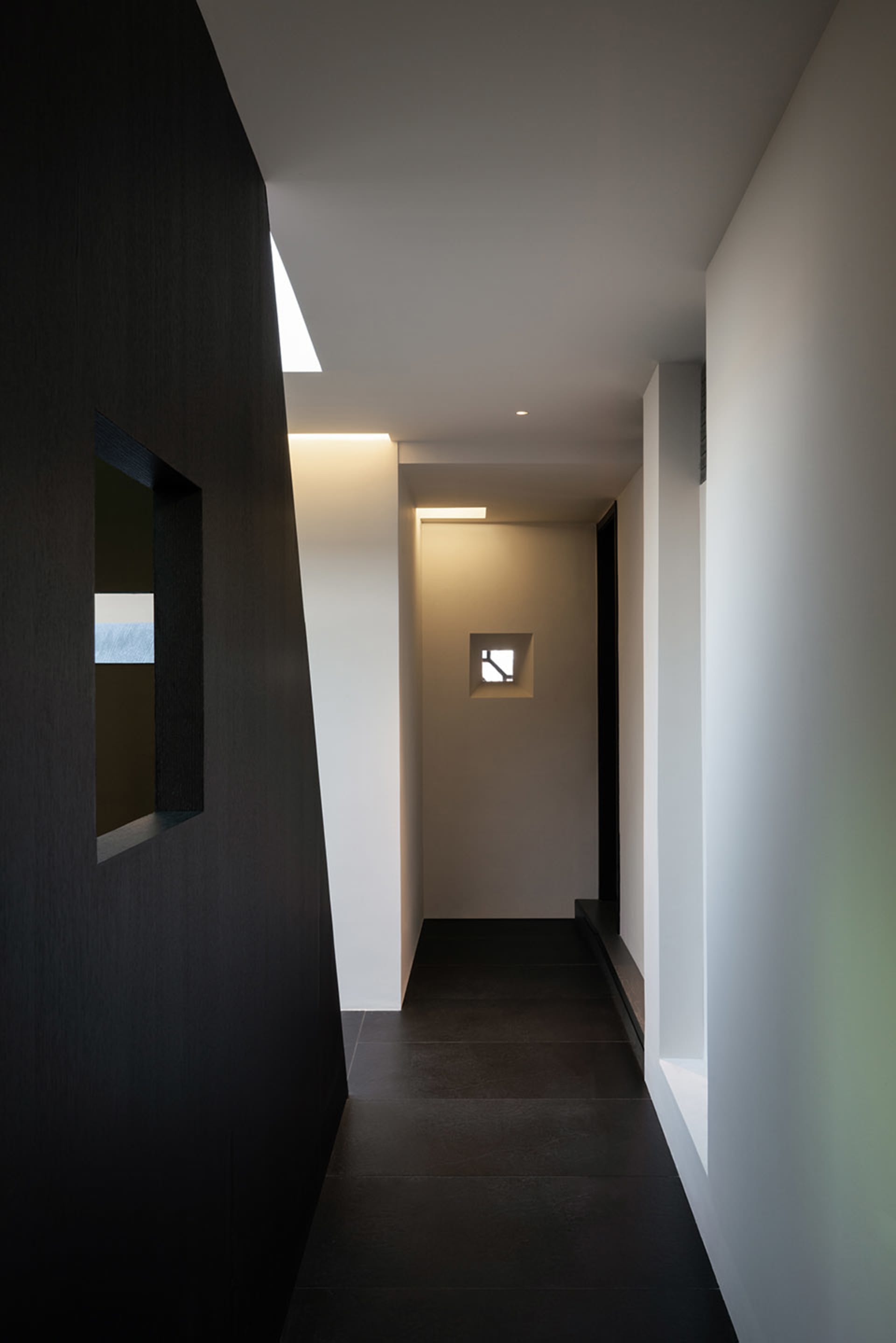 Narrow hallway with a black door and two windows on a white wall, creating a somewhat dark atmosphere