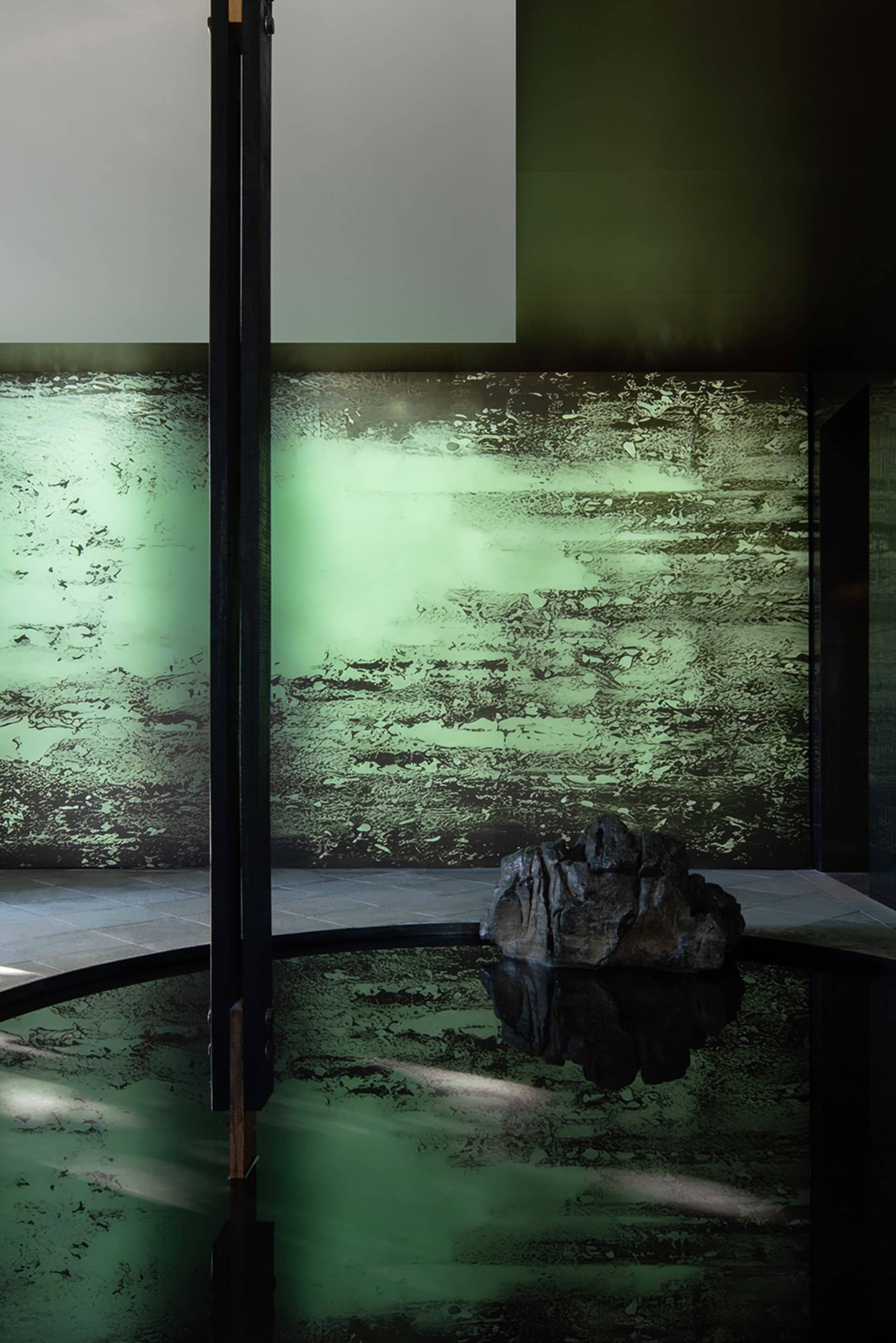 A modern pool area with a large rock in the pool, surrounded by a green wall and two black poles, with a sleek and modern design
