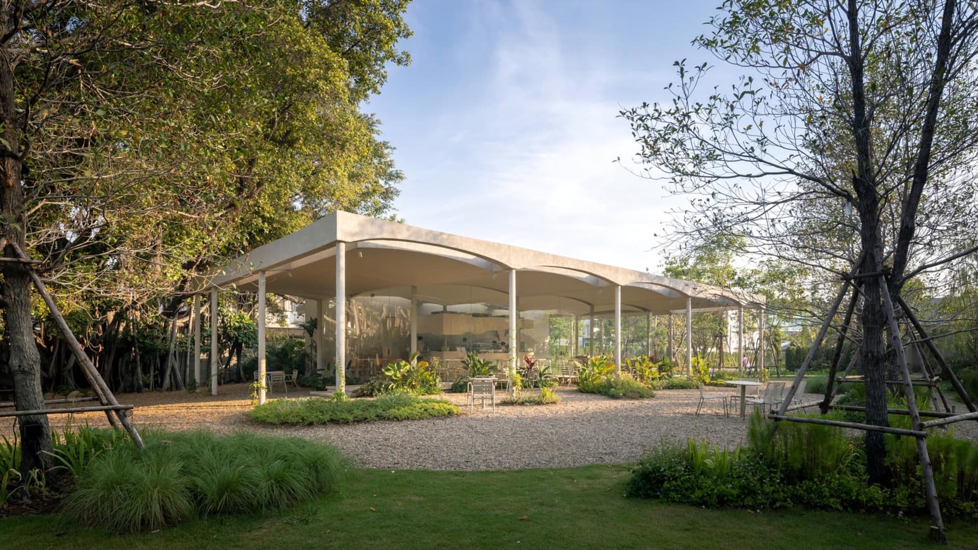 An open air living area with a large white structure featuring a glass roof, multiple rooms, chairs, and potted plants, creating a relaxed and inviting atmosphere