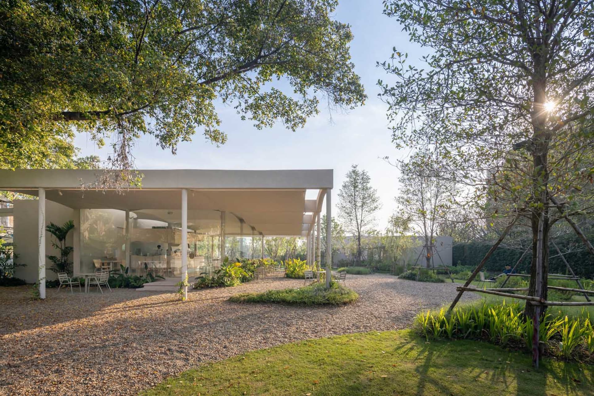 Open-air living area with a modern building featuring glass walls, potted plants, and chairs, blending indoor and outdoor spaces