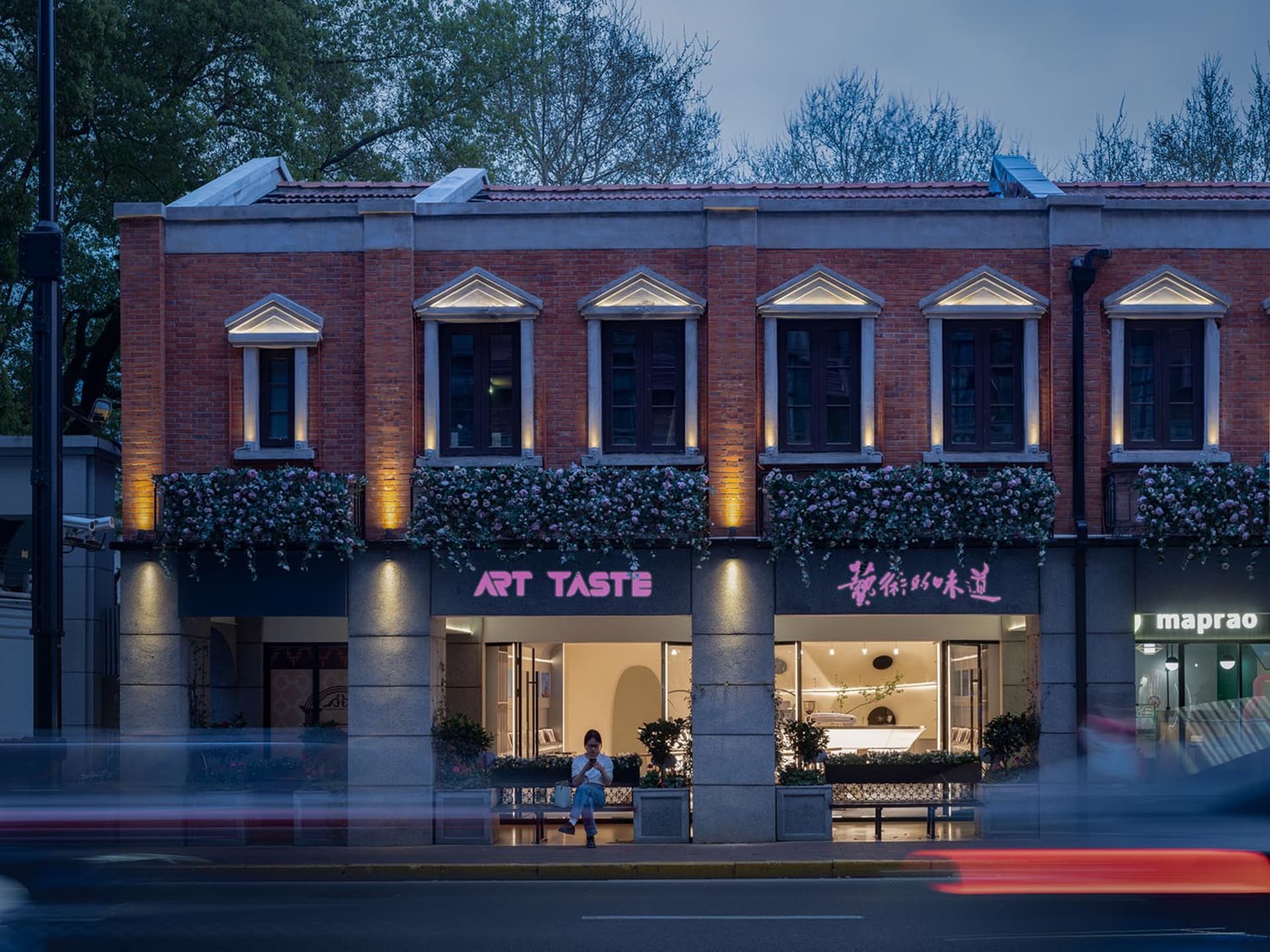 A brick storefront at night with a pink sign, potted plants, and a busy street scene in the background