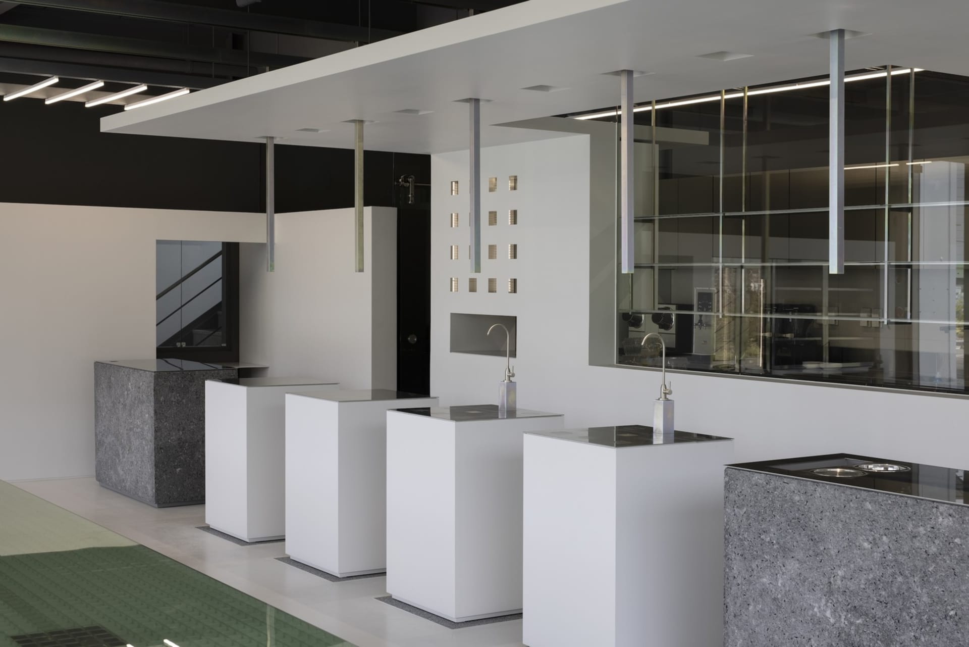 A modern bathroom with white fixtures, three sinks, two toilets, and a large glass wall, giving a clean and minimalist impression with natural light