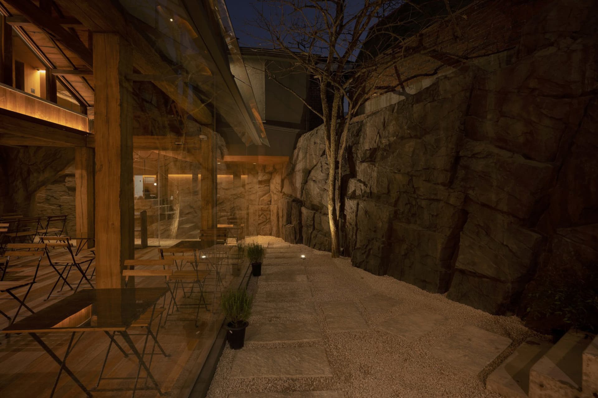 夜晚戶外餐區，桌椅與盆栽置於照明建築前，碎石步道旁有石牆