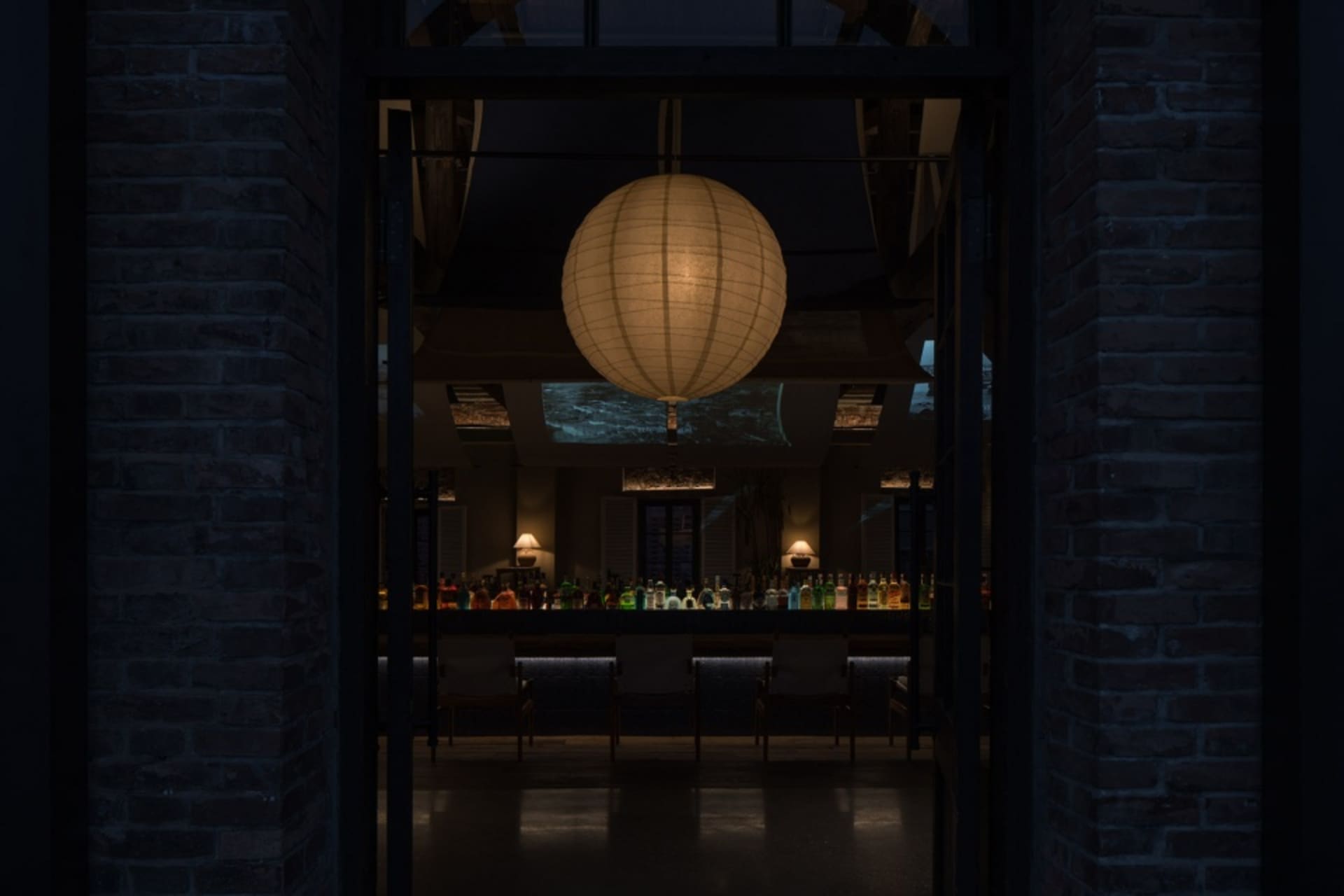 A cozy restaurant with a round lamp as a centerpiece, surrounded by chairs and dining tables, with bottles on surfaces throughout