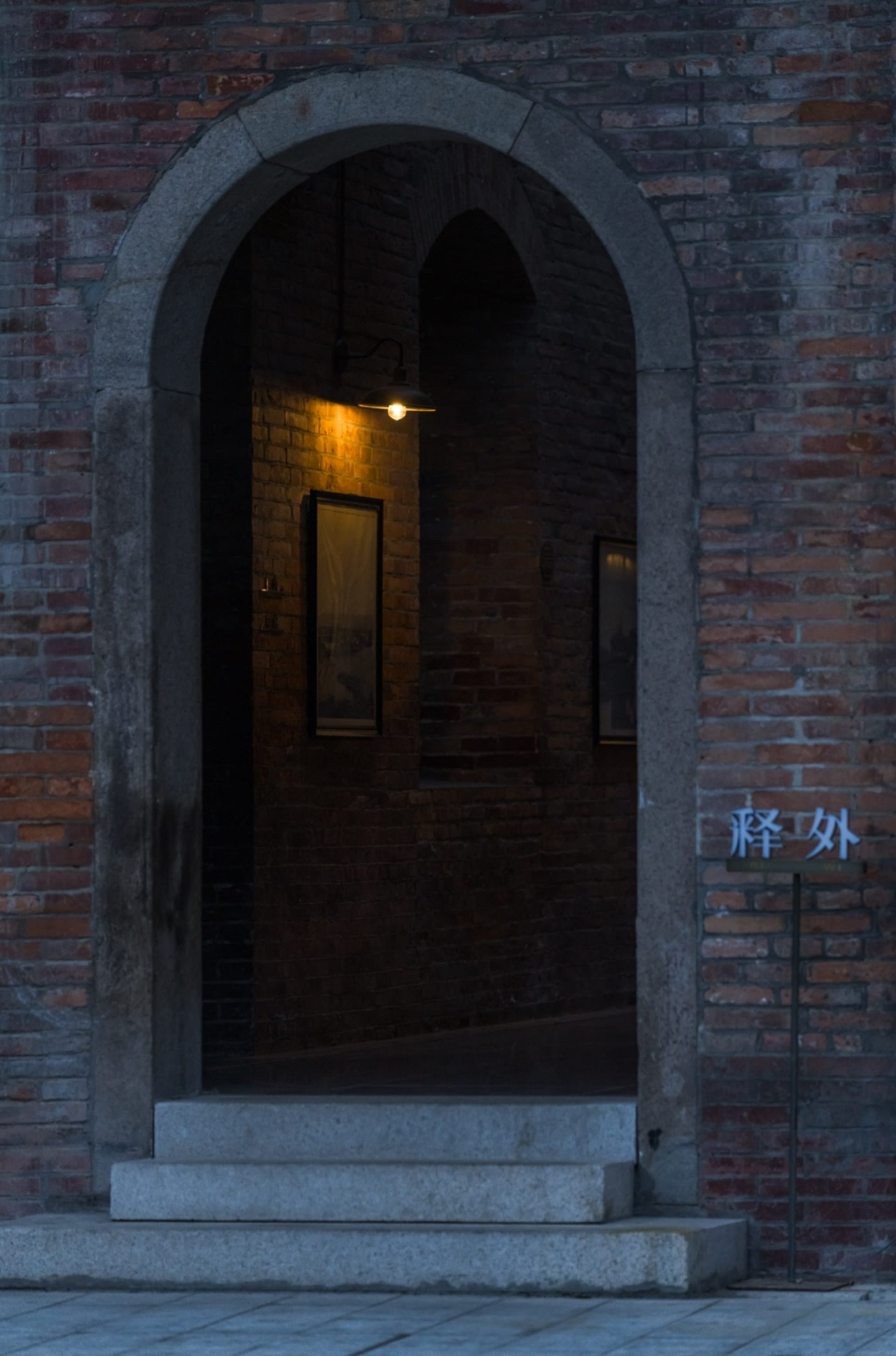 Dimly lit hallway with a brick wall, two framed pictures, and an archway leading to a dark space