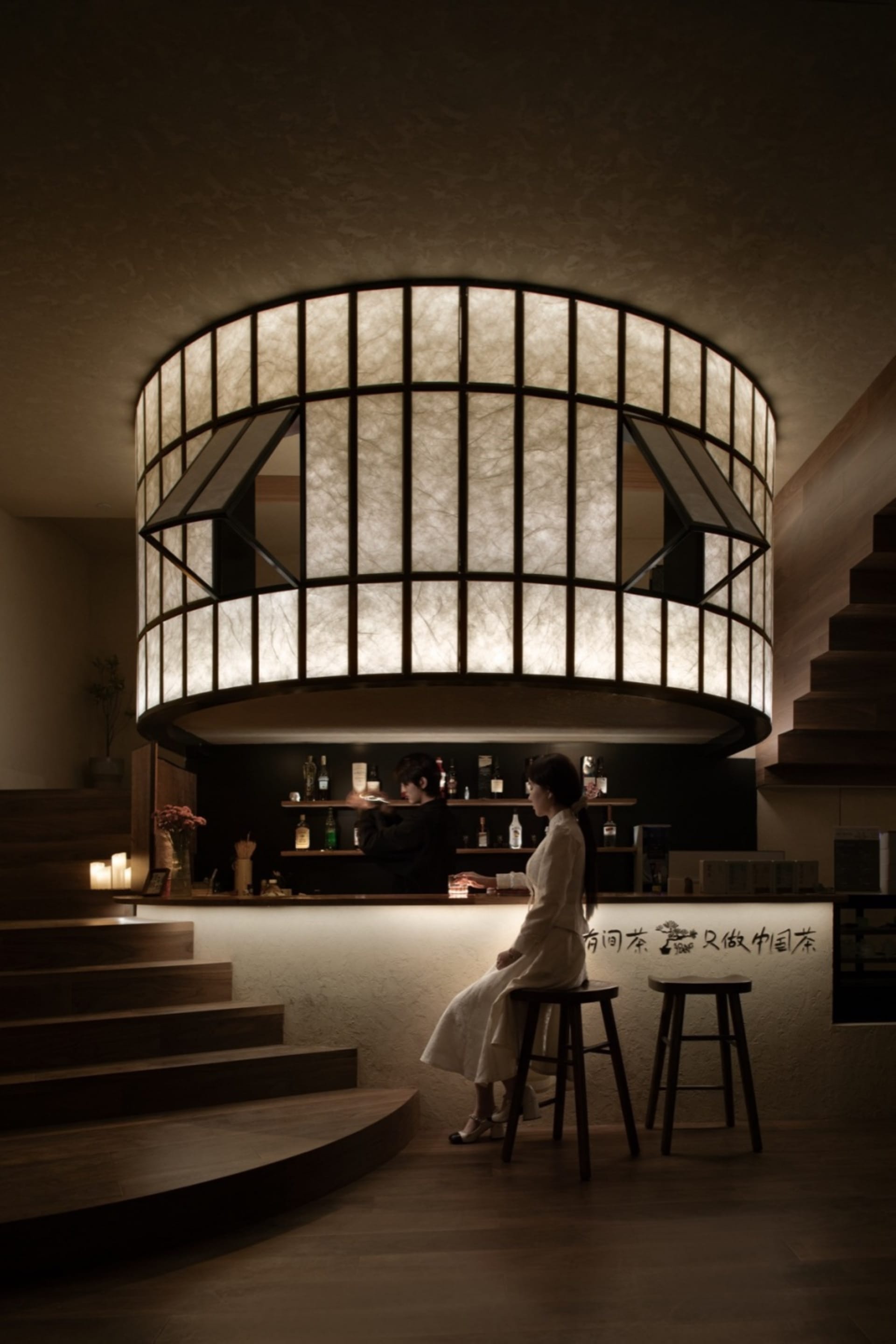 A woman sits at a counter in a relaxed Asian bar with bottles and cups scattered around her