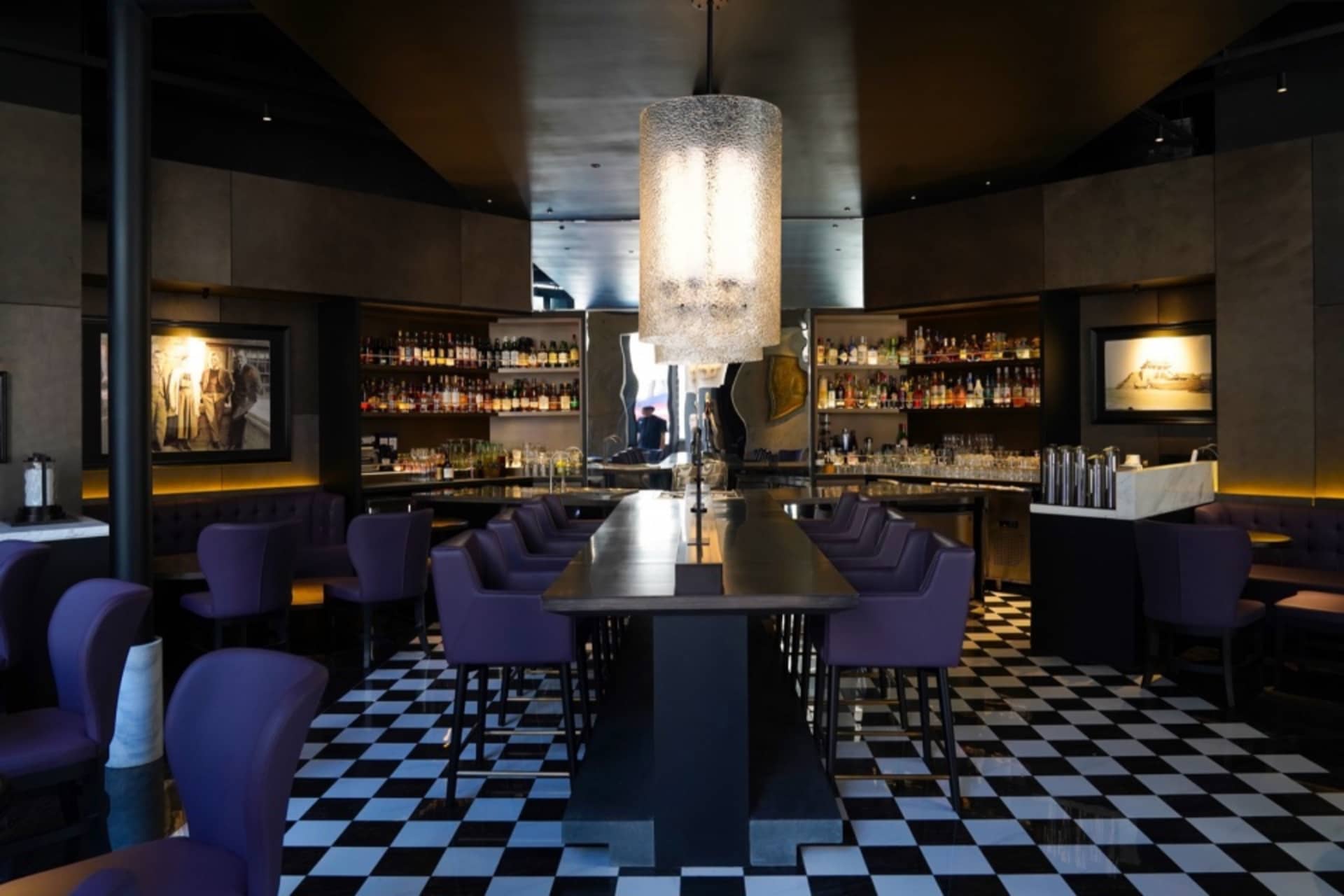 A modern bar area with a long dining table, black and white checkered flooring, and several bottles on the table