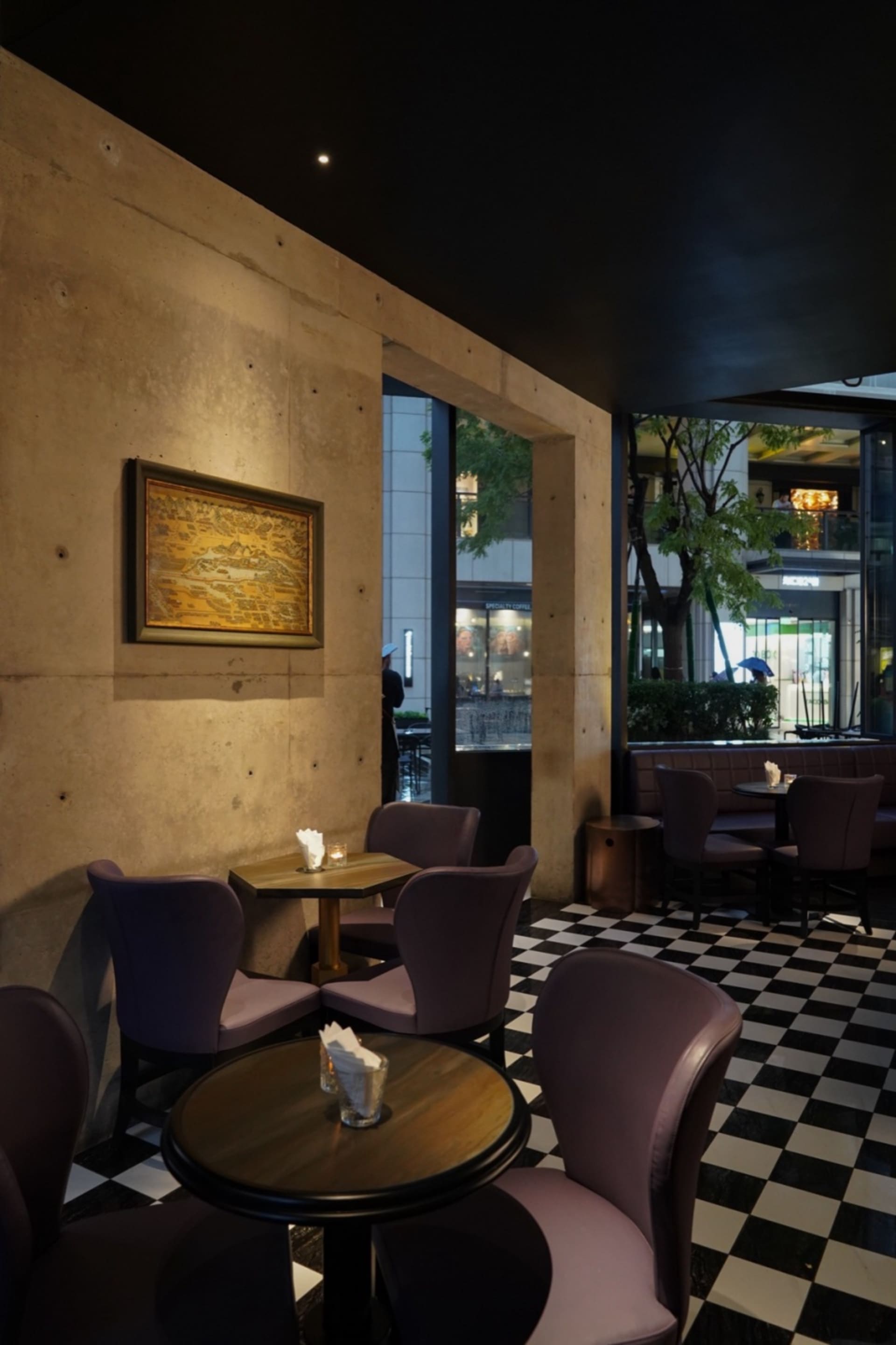 A modern restaurant with a large dining table and several smaller tables, purple accents, and a concrete finish, lit by lamps creating a warm ambiance