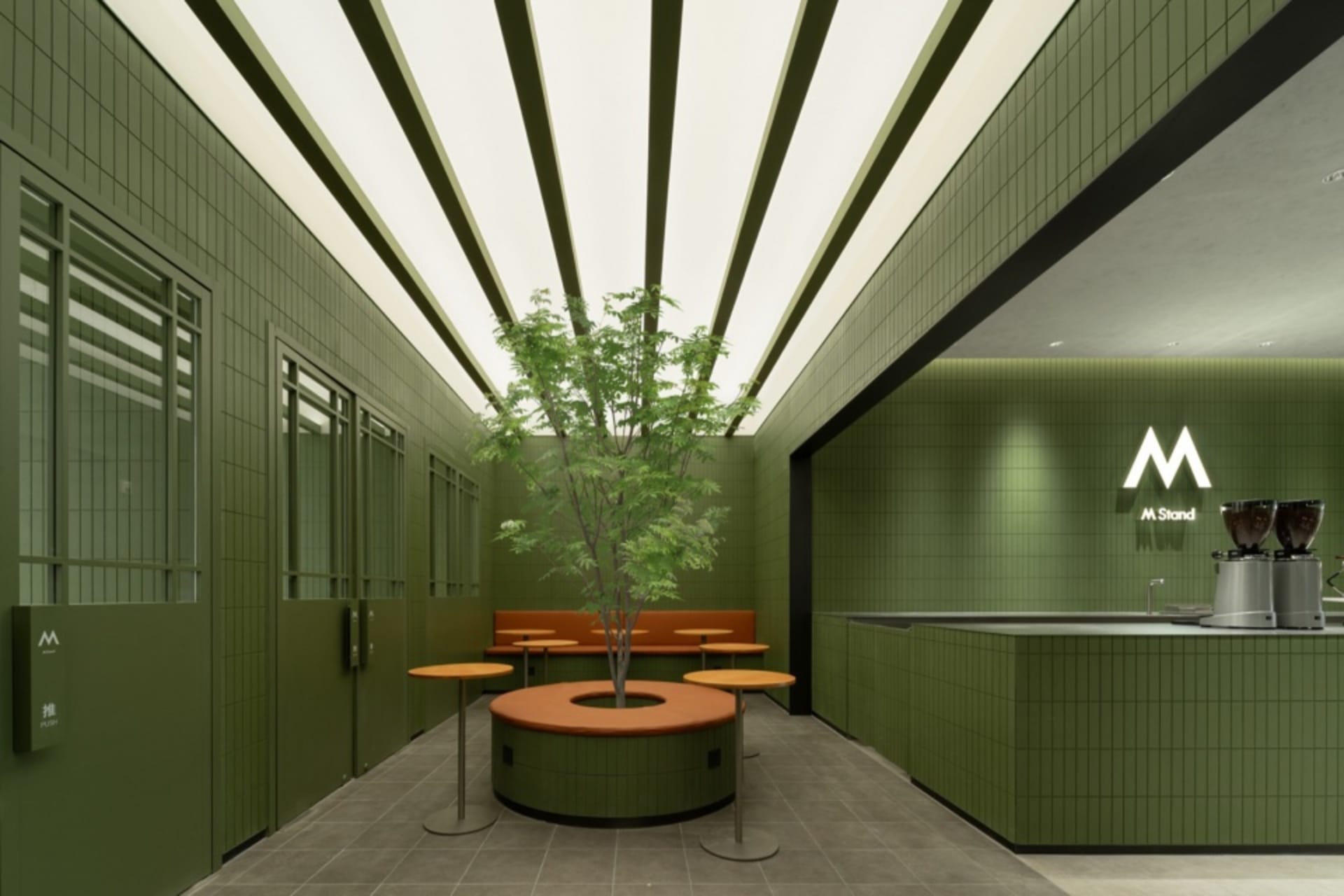 A restaurant interior with green walls, a large tree in a pot, and dining tables with chairs, creating a welcoming atmosphere with a focus on nature and greenery