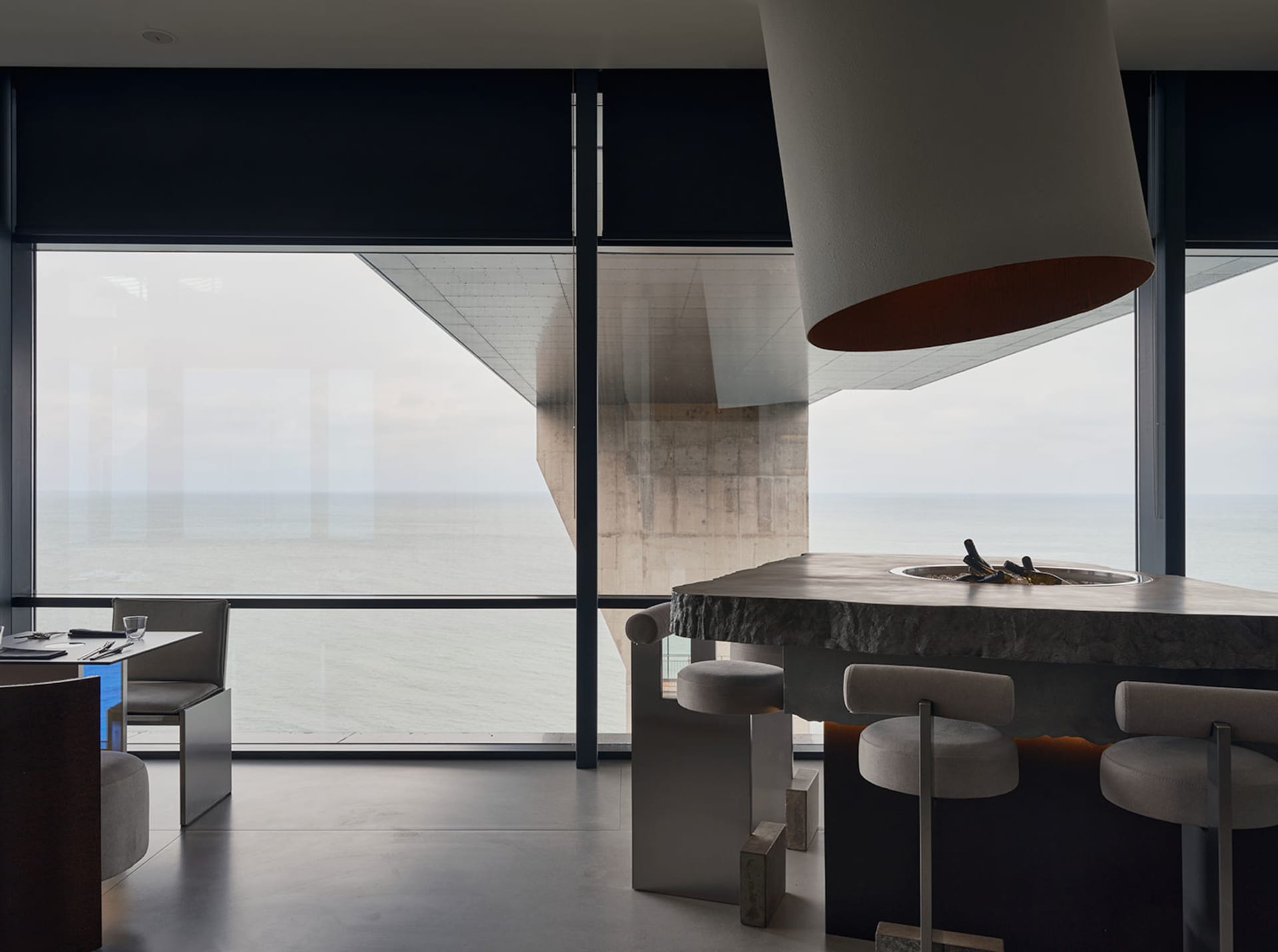 Open modern living space with a kitchen island, stools, chairs, and a large window providing natural light, featuring a marble countertop