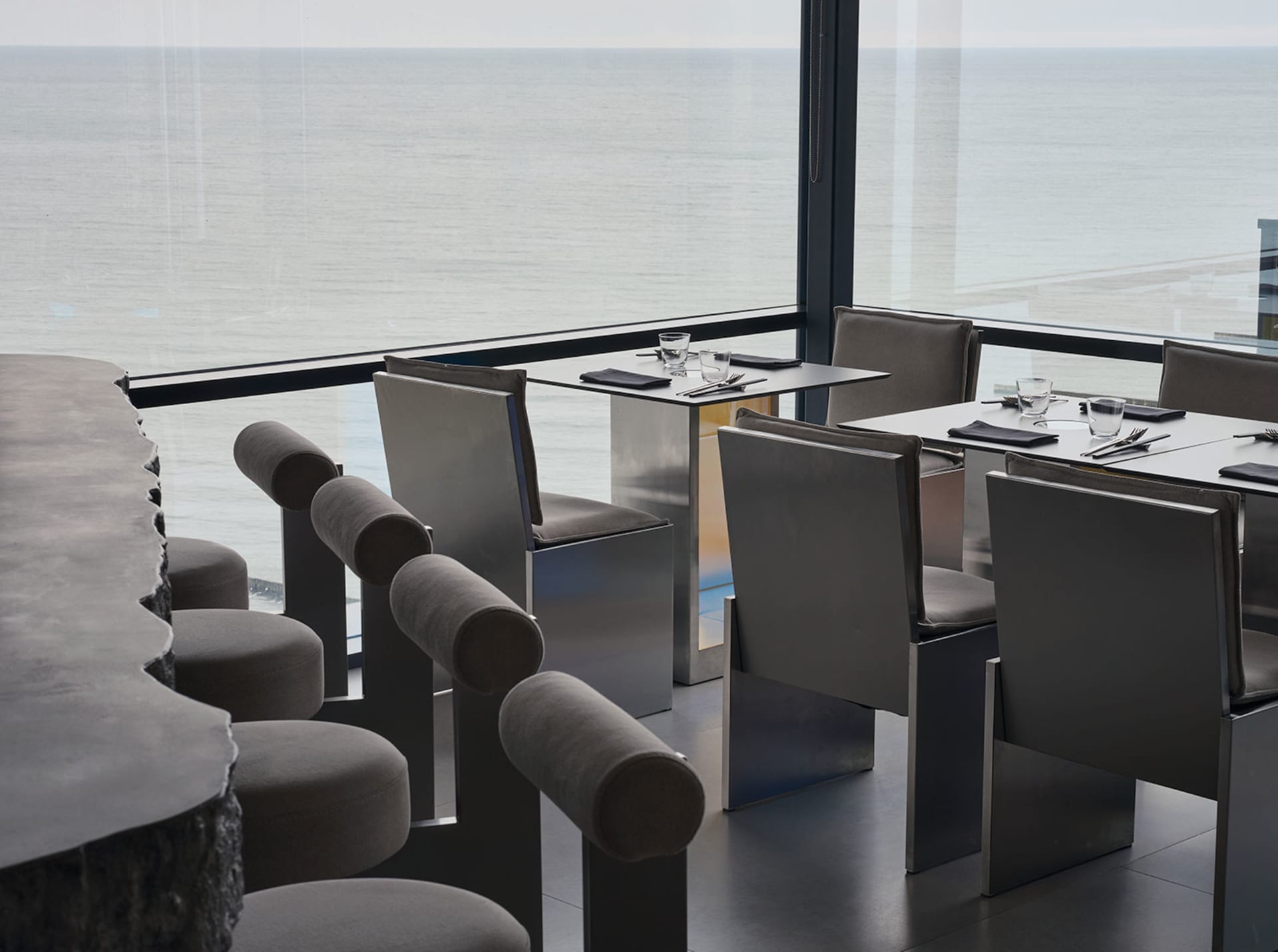 Sophisticated dining area with a large glass window offering a stunning ocean view, surrounded by chairs, a dining table set with wine glasses, cups, cutlery, and bottles