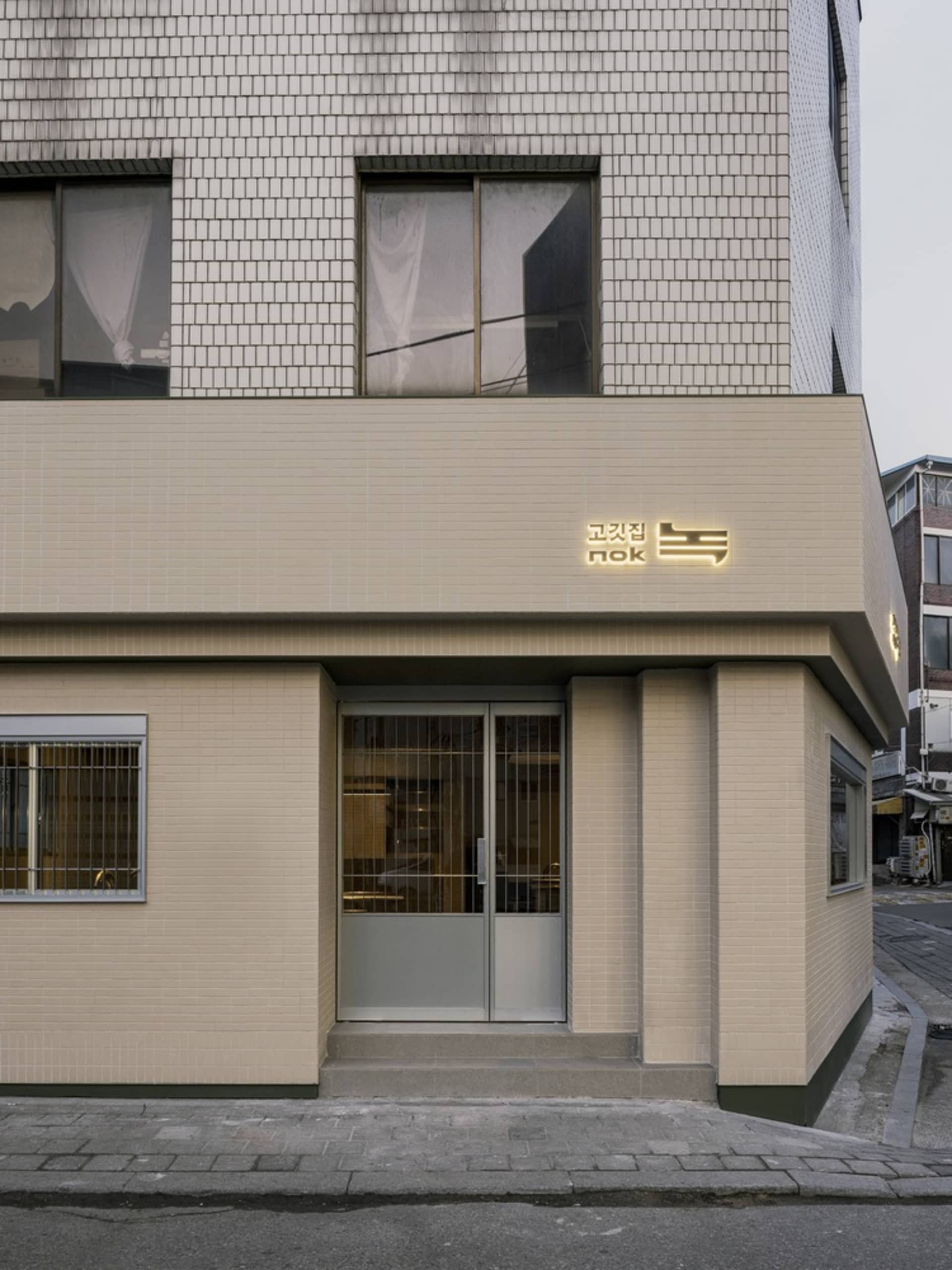 磚牆建築搭配白色立面與大片窗戶，背後為都市景觀，外頭停有一輛車