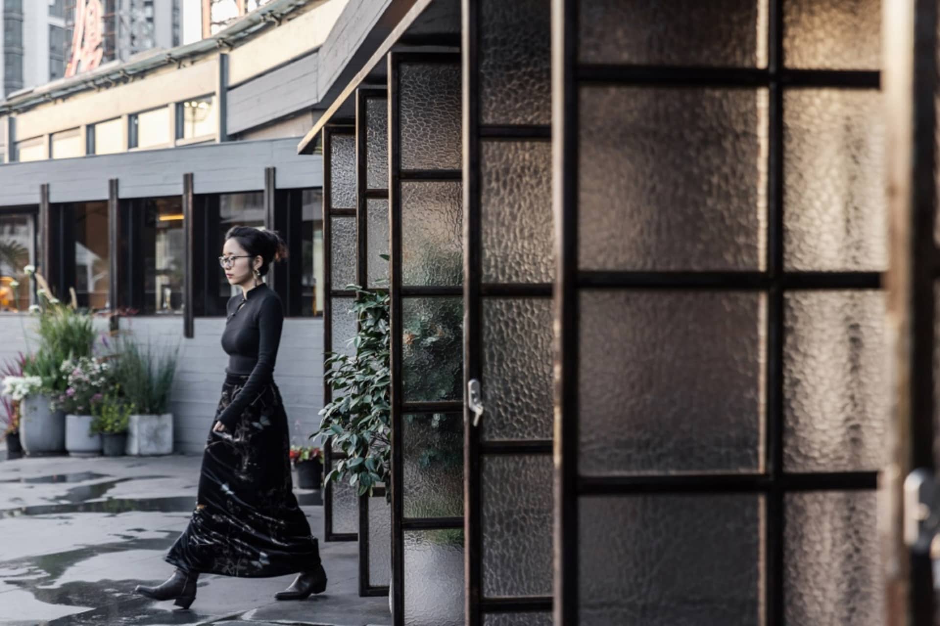 An Asian woman walks past a potted plant in front of a restaurant with a glass door, wearing a black dress and glasses