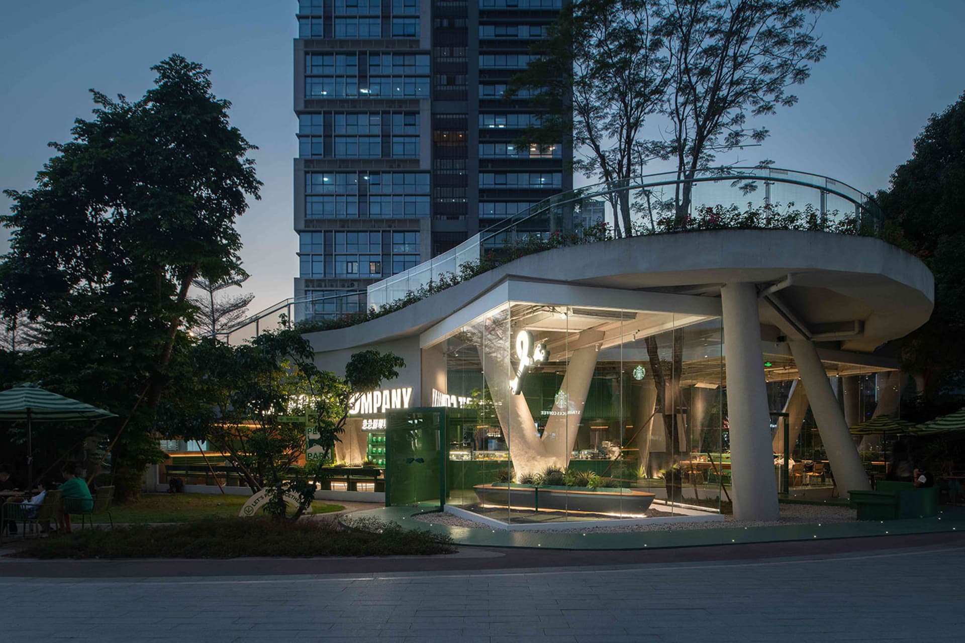 A modern building at night with a large glass structure, green roof, and rooftop terrace, surrounded by trees and potted plants