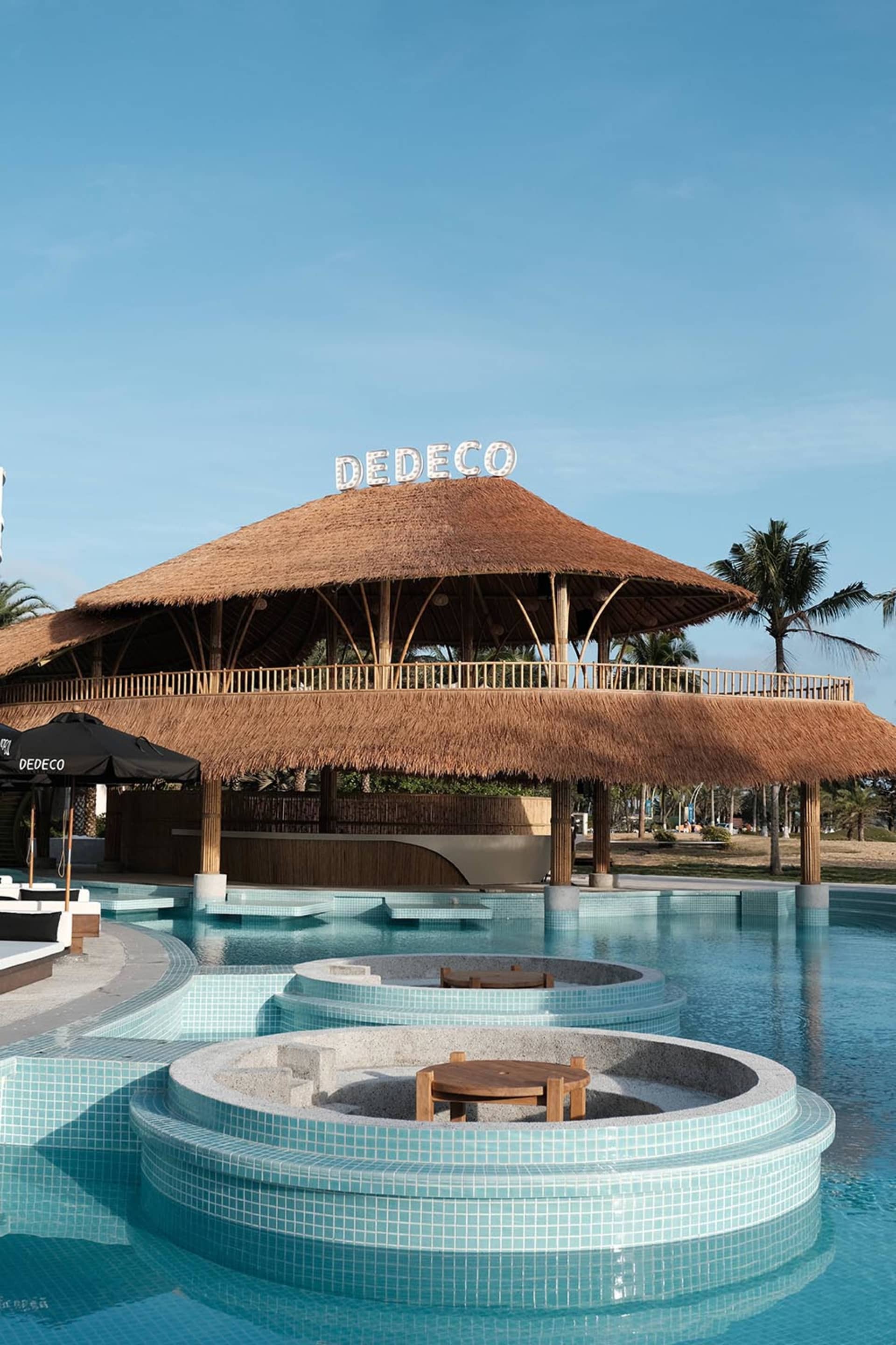 A serene outdoor pool area with a thatched roof, chairs, and a view of the water