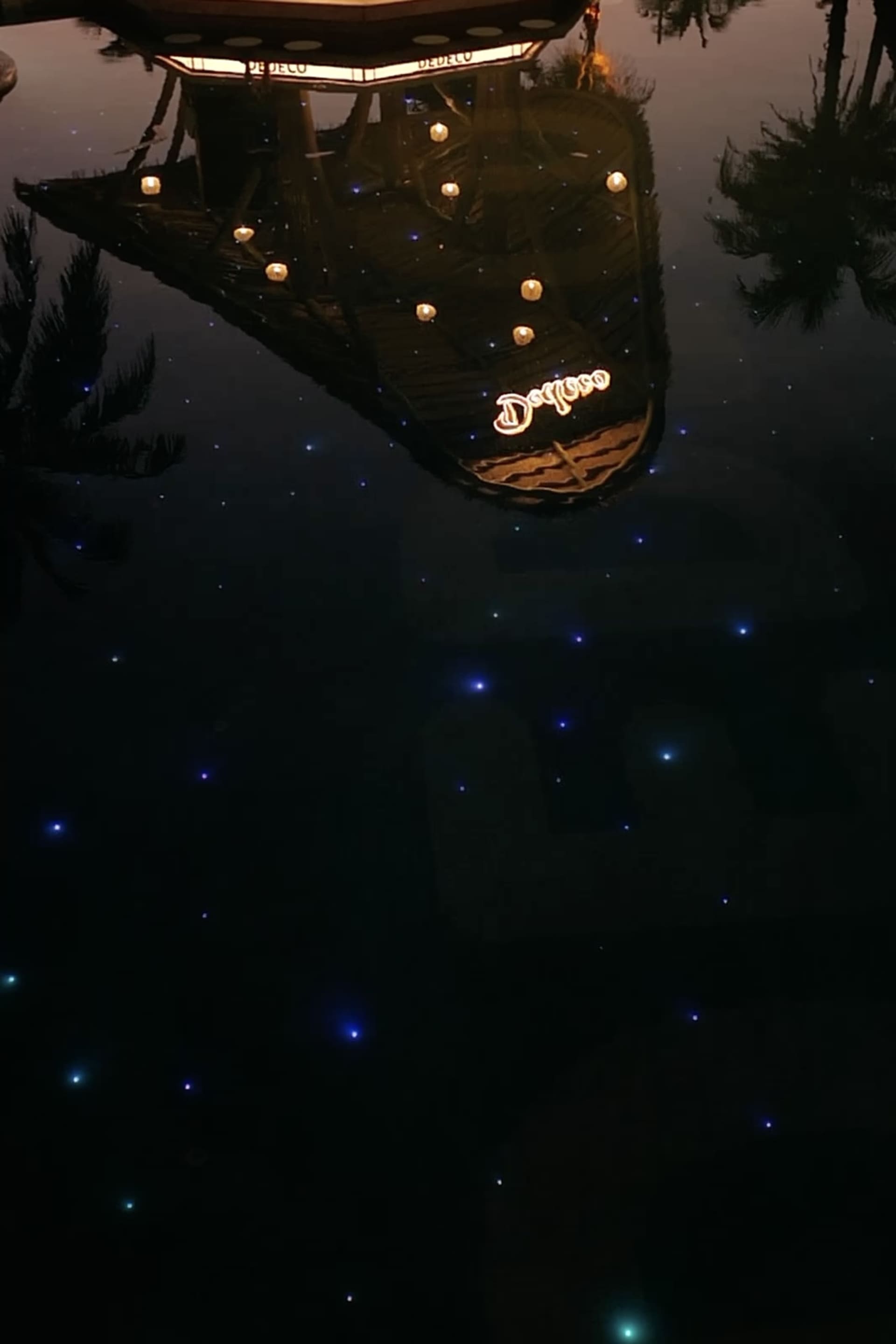 A wooden boat on water at night surrounded by stars, illuminated by numerous lights