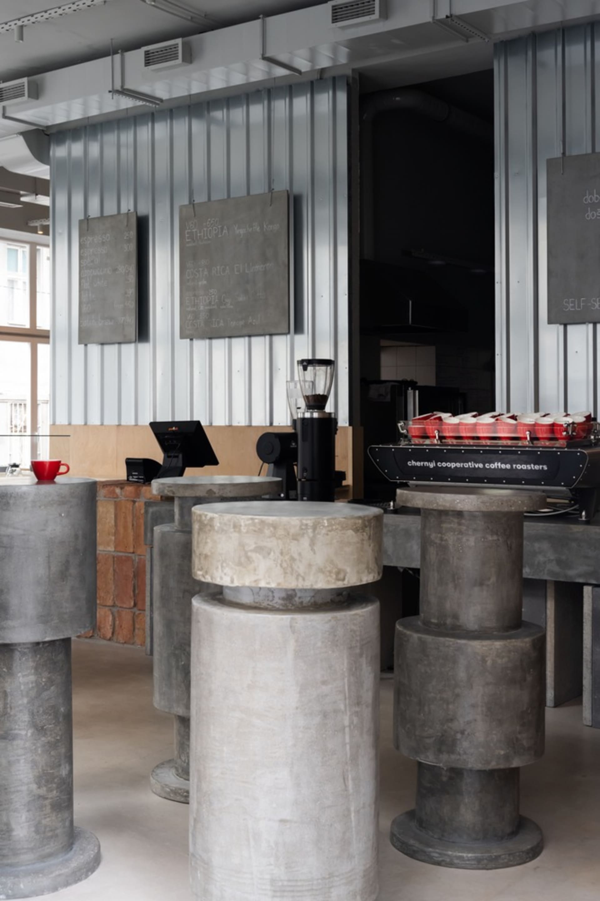 Industrial coffee shop with concrete walls, pillars, and blocks, featuring a dining area with chairs and a refrigerator, and a cozy atmosphere with various decorative items