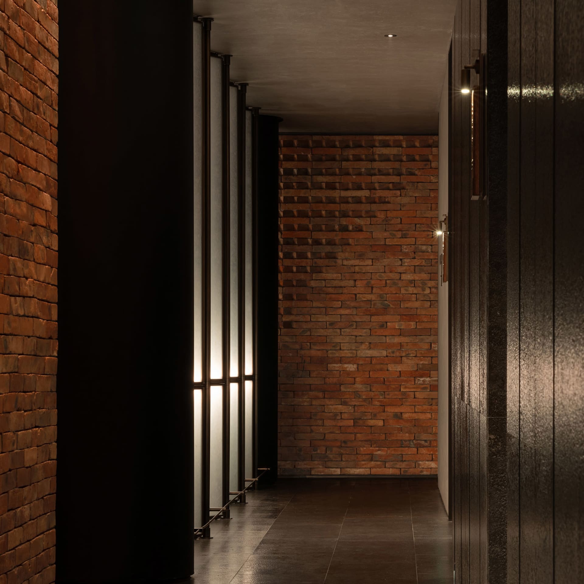 A dimly lit hallway with red brick walls, black and brown doors, and two windows providing natural light