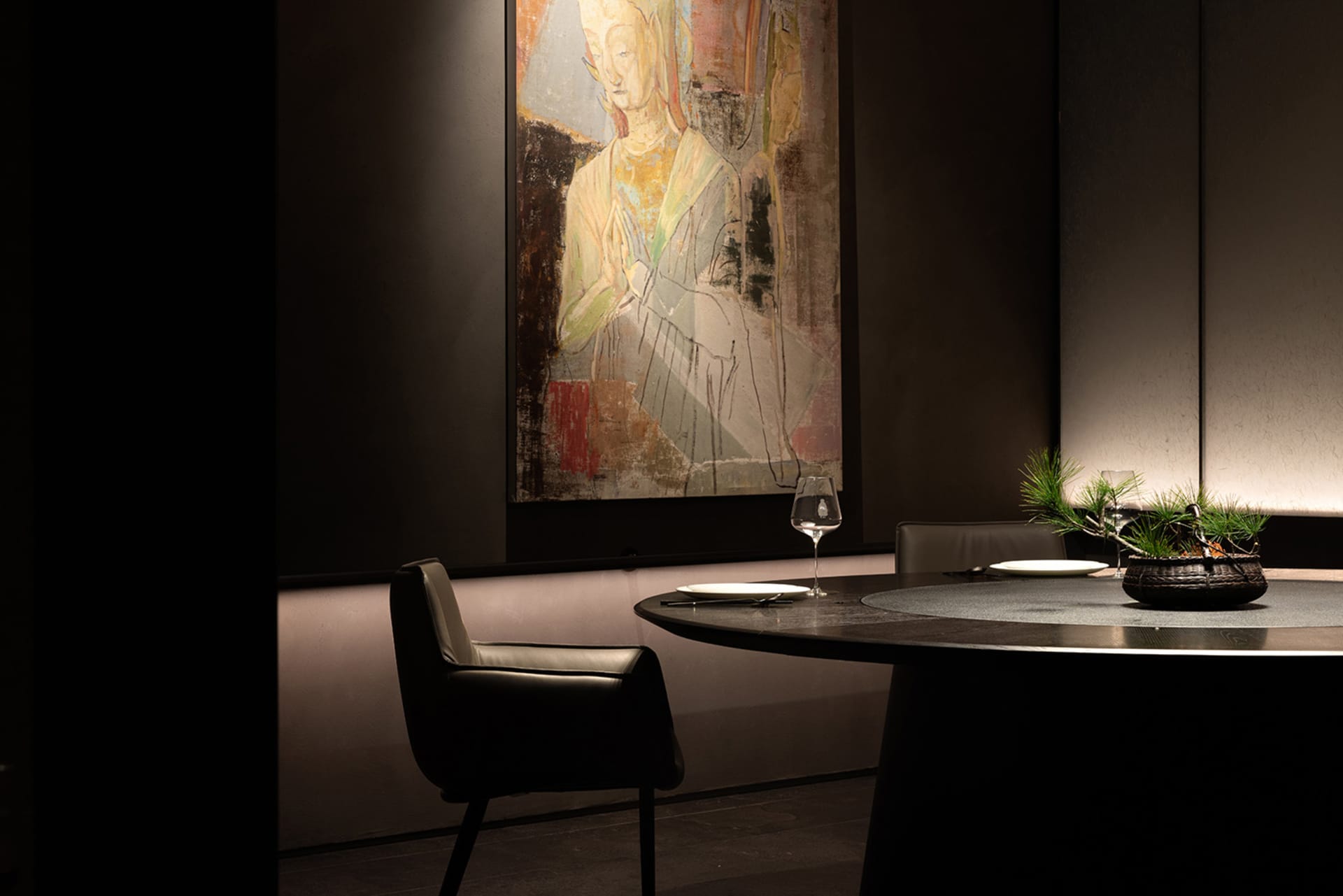 A woman's portrait in a black frame on a wall, with a dining table and chairs, wine glasses, and a potted plant in the corner