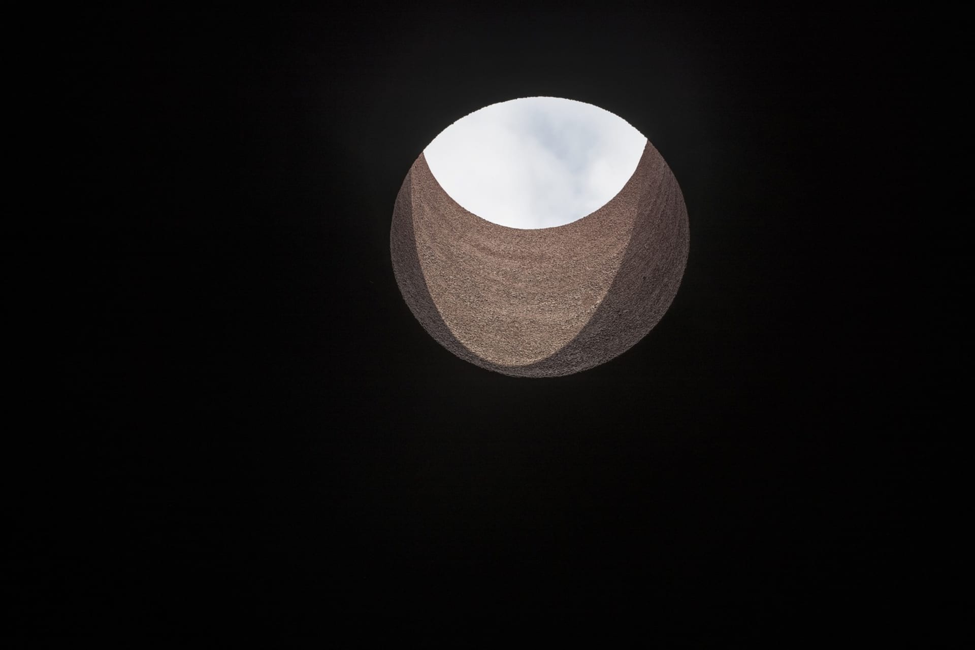 A modern indoor scene featuring a round hole in a wall, surrounded by concrete or stone walls, with black and white colors and overhead lighting