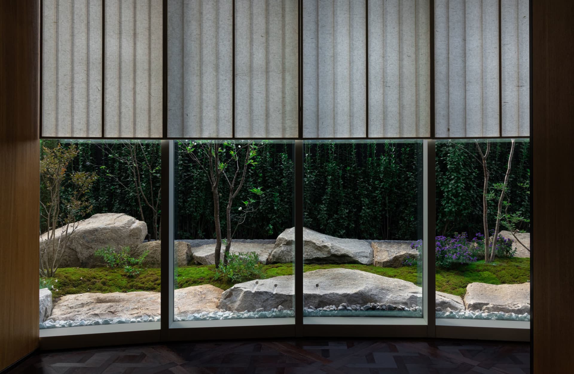 A tranquil indoor space with a large window offering a view of a garden with a pond, rocks, and plants, surrounded by trees, and natural light pouring in