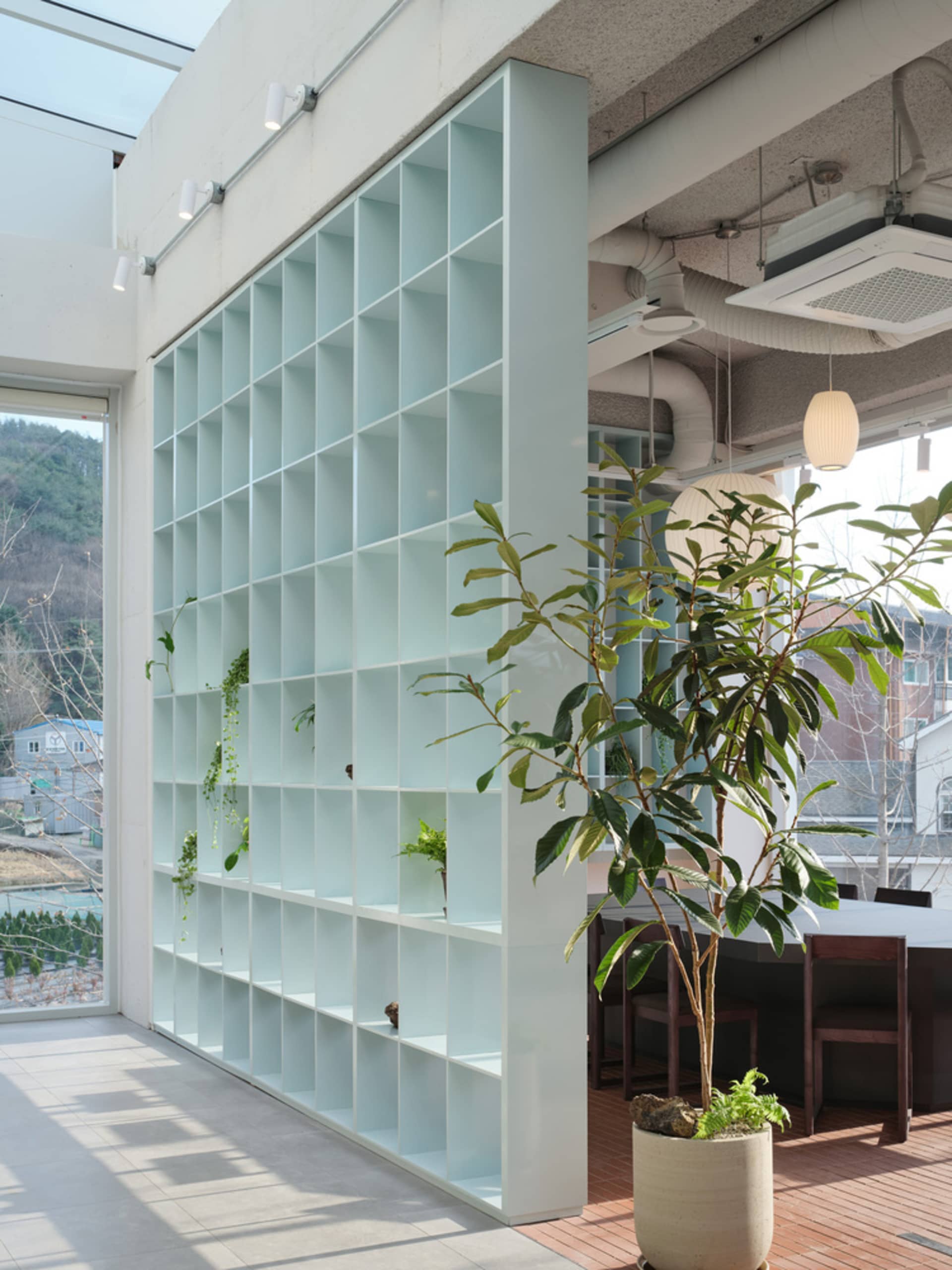 A modern living or working space with a glass wall, bookshelves, chairs, and a dining table in a well-lit environment