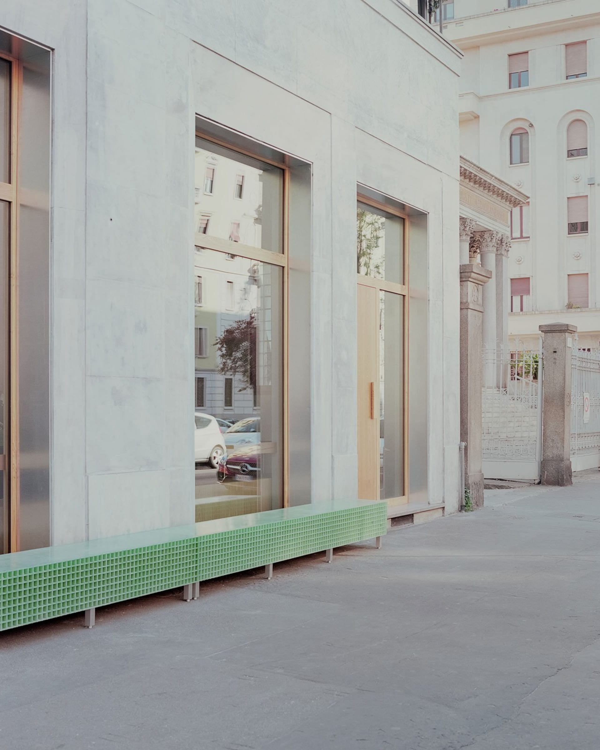 A modern building with a metal bench in front of its windows, surrounded by parked cars in an urban setting
