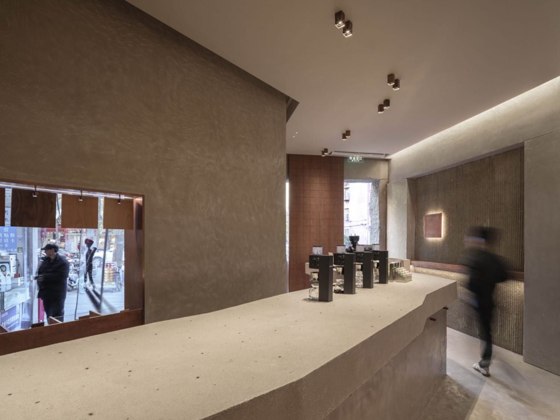 A casual cafe interior with a counter area, wooden accents, and a person walking past, serving beverages to customers