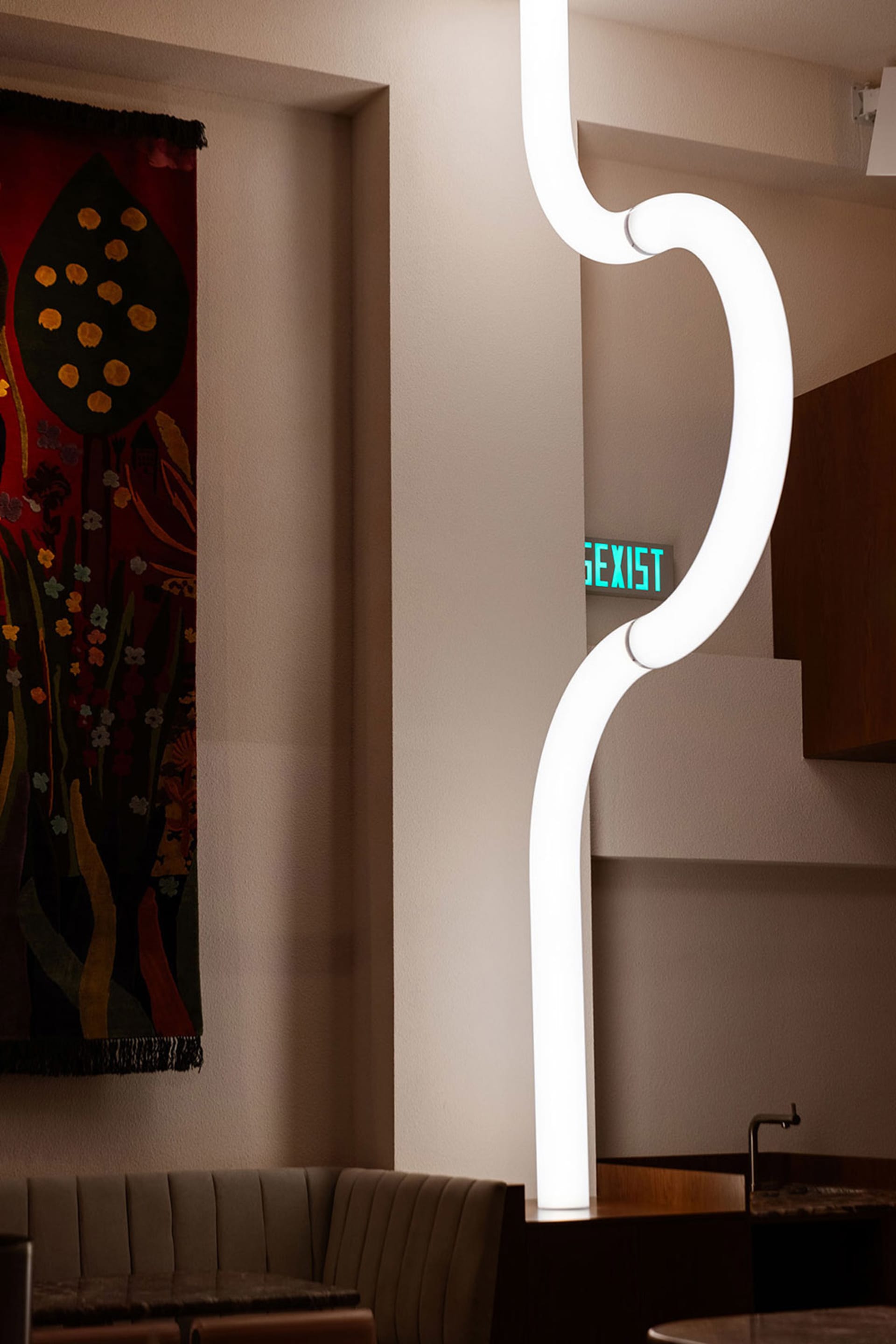 A modern room with a large white wall featuring a green sign reading sexist, a curved light, and a painting on the wall