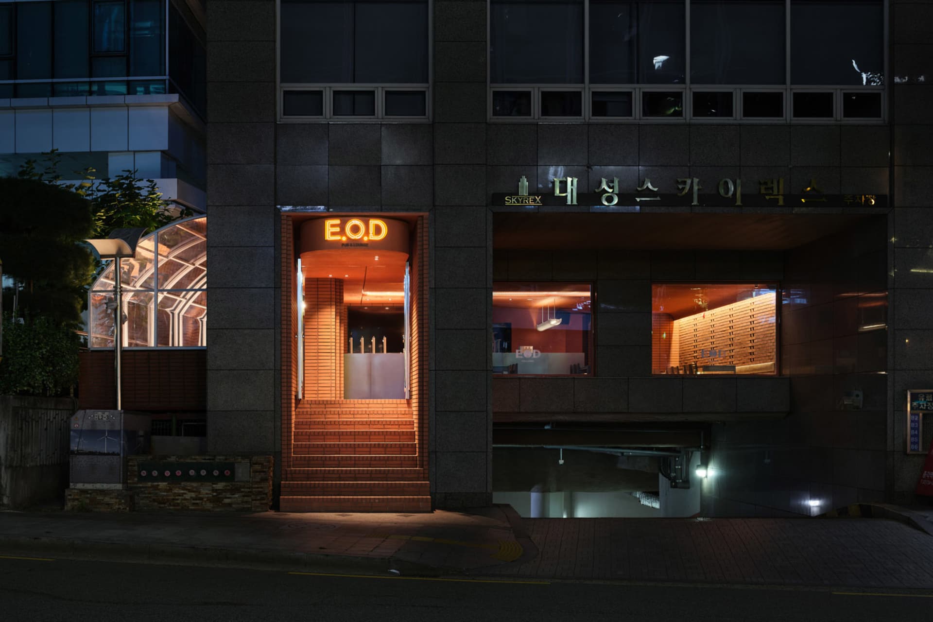 夜晚現代亞洲餐廳，磚牆外觀、玻璃窗，溫暖室內氛圍，設有餐桌、椅子與酒瓶